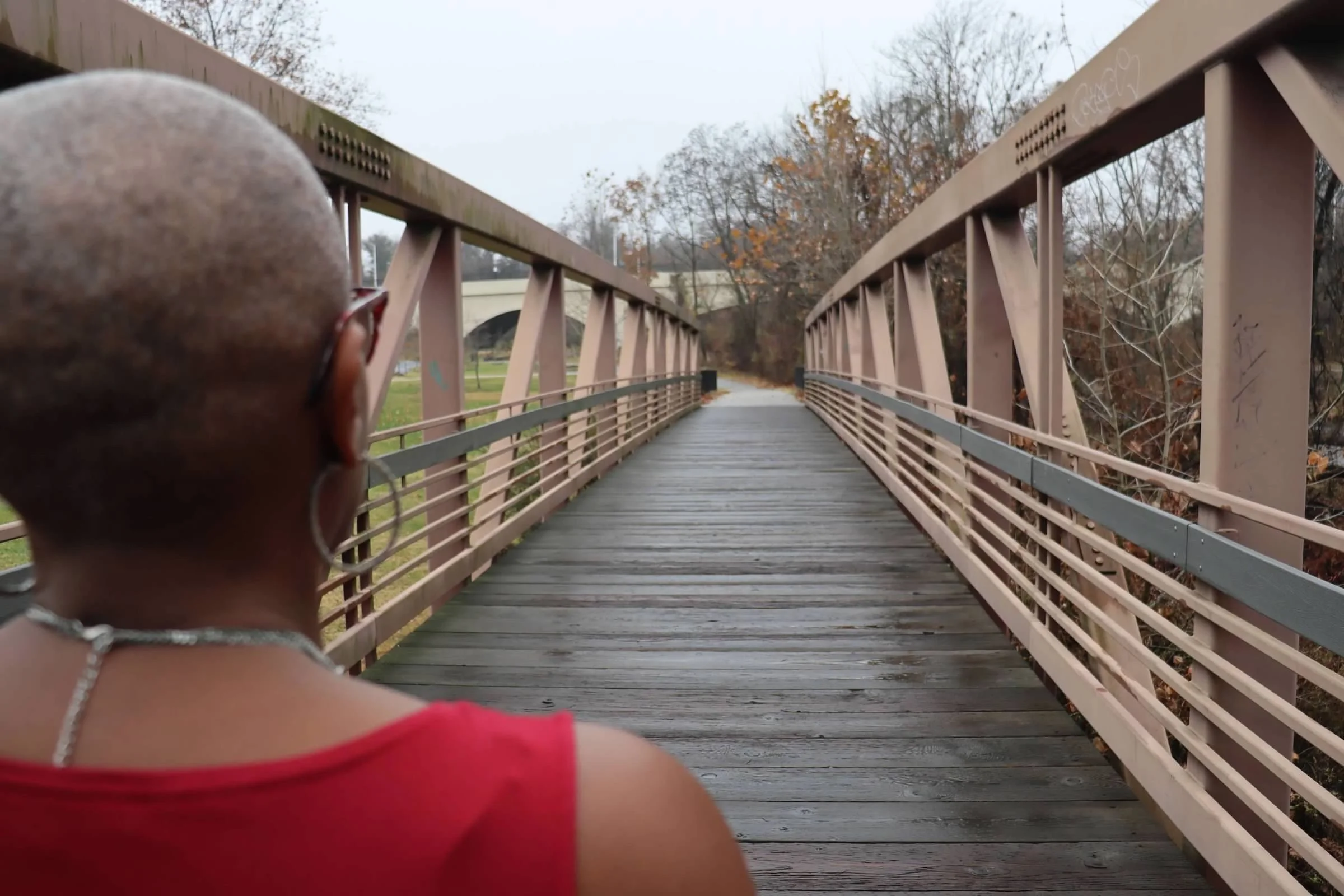 Person with a shaved head and earrings standing on a wooden bridge during daytime.