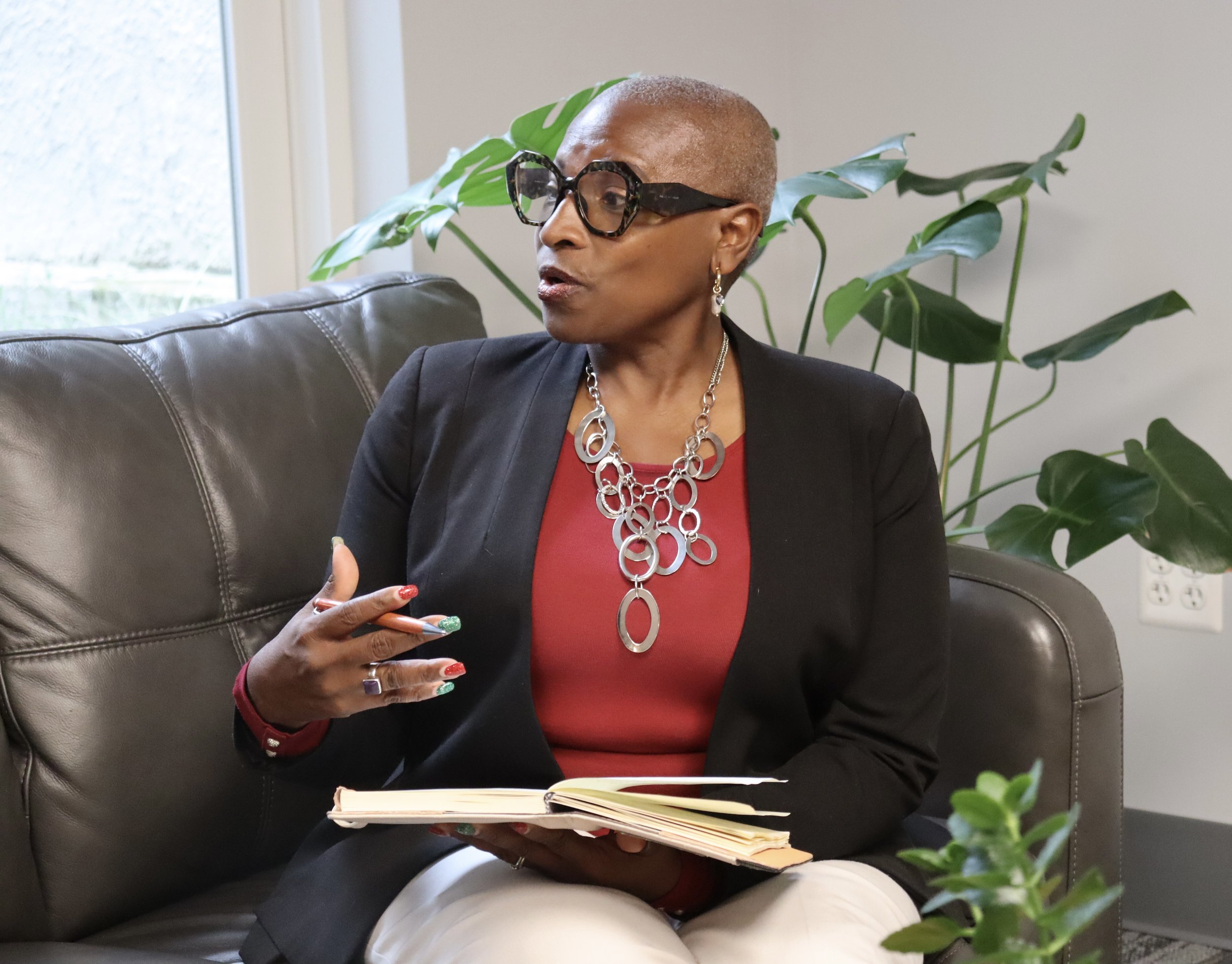 An older woman with short, gray hair, wearing large black glasses, gold earrings, a silver jewelry necklace, a black blazer, and a red top, sitting on a black leather couch next to a large green plant, holding a smartphone in one hand and a notebook in her lap, with a window and white wall in the background.