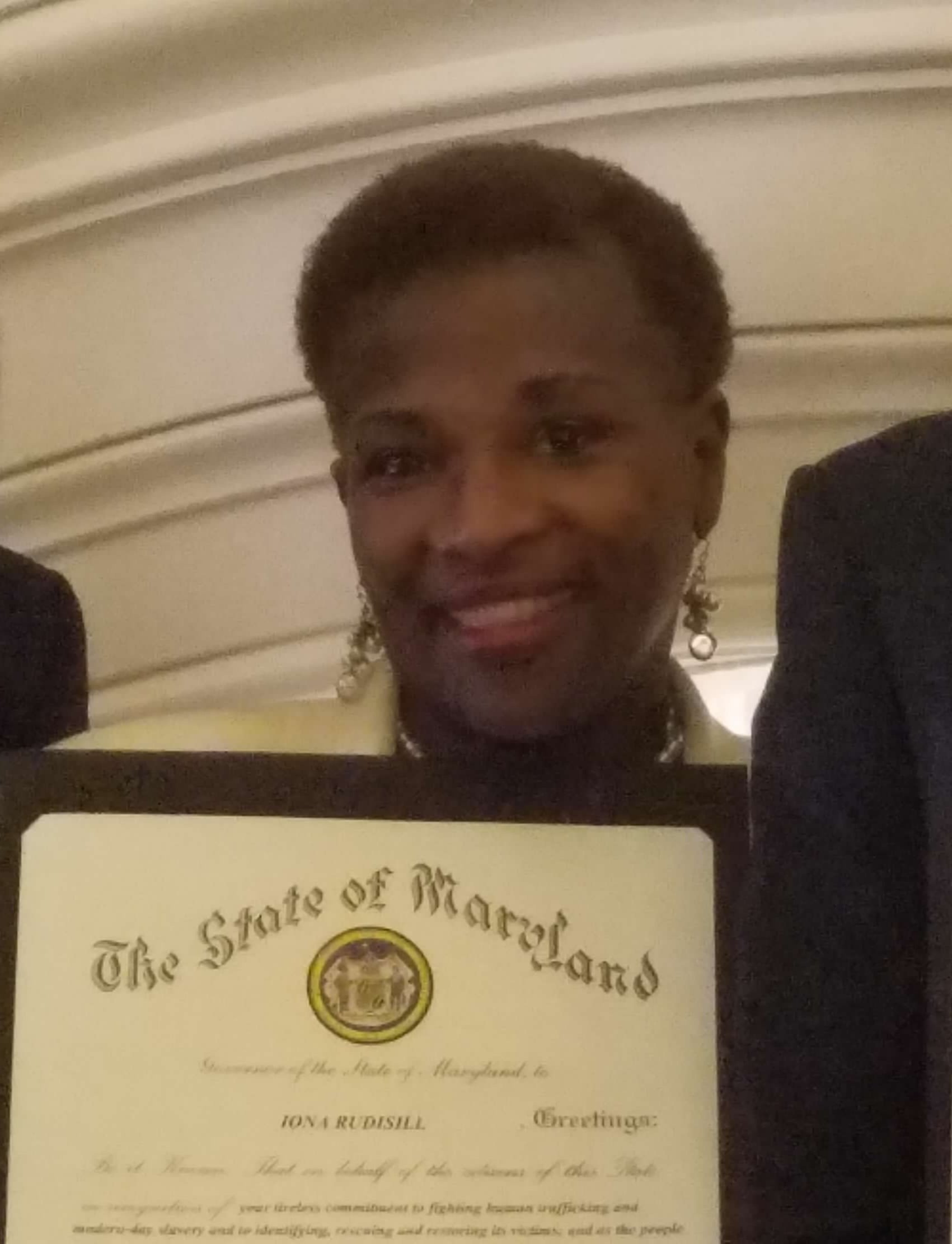 Part of a certificate titled 'The State of Maryland' with a seal and text, held by a smiling person with short hair, earrings, and dark clothing, in front of a beige wall with decorative molding.