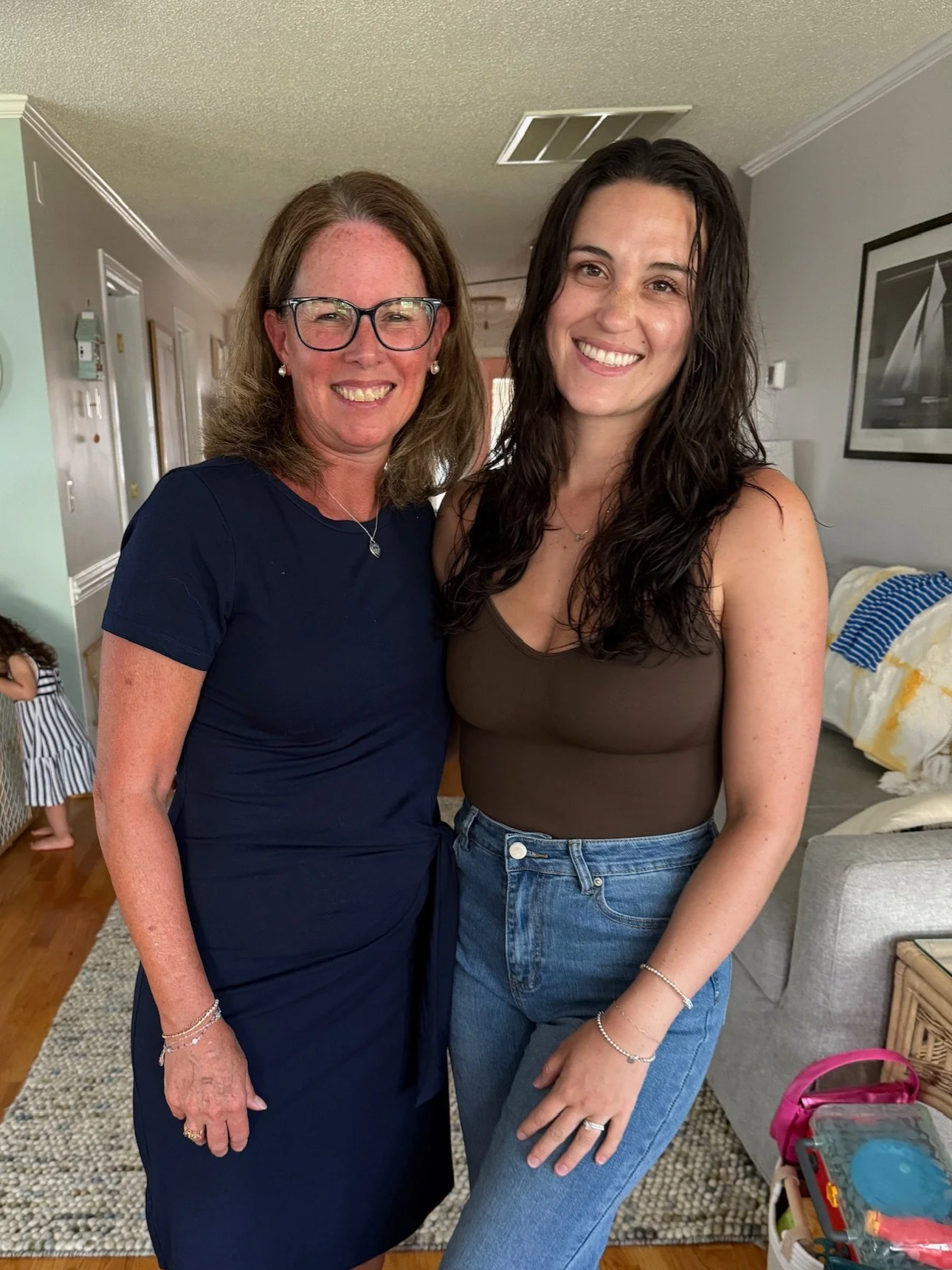 Two women smiling and posing inside a home, with a child partially visible in the background.