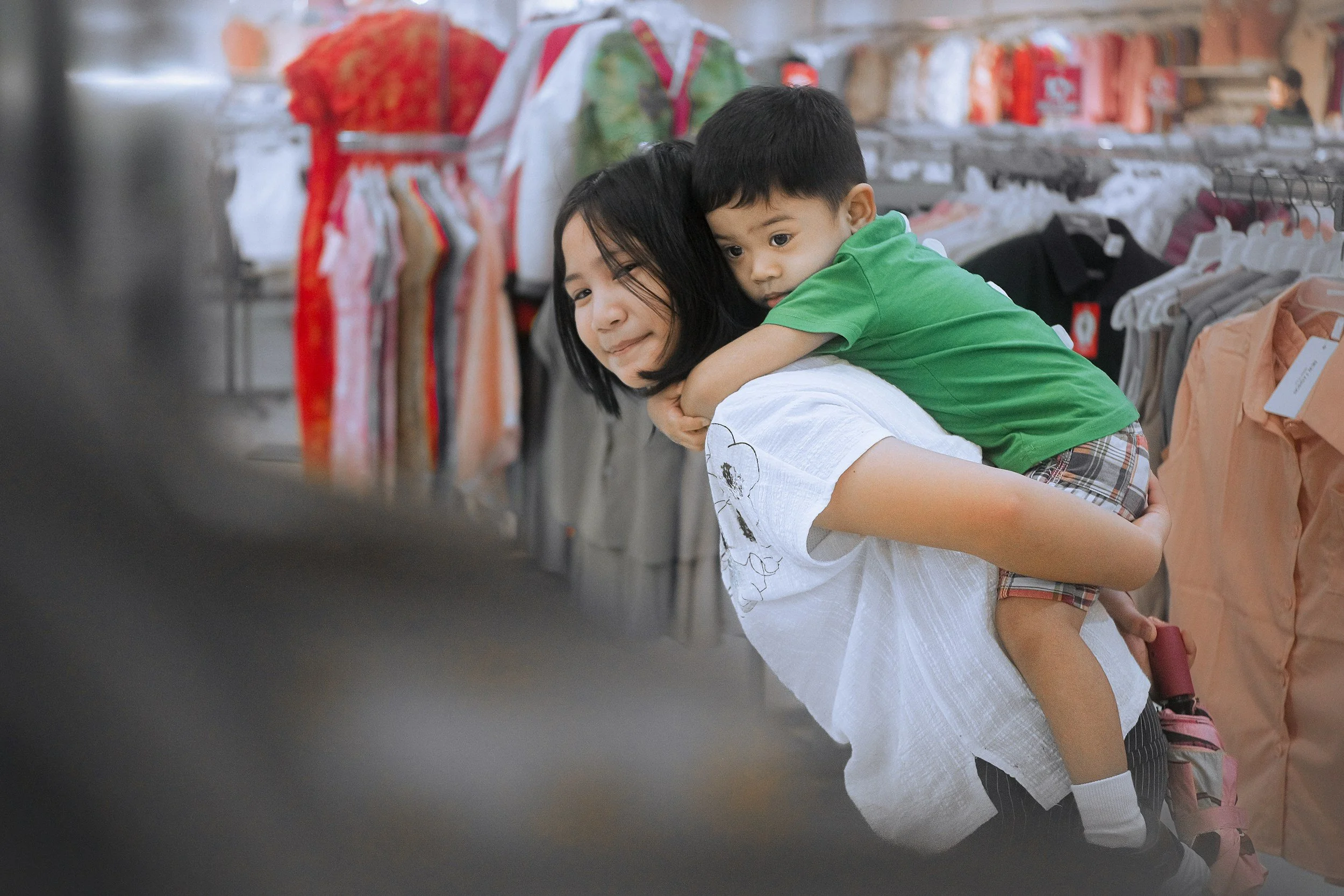 A young woman with shoulder-length dark hair carrying a small boy on her shoulders in a retail store aisle, with racks of clothing in the background.