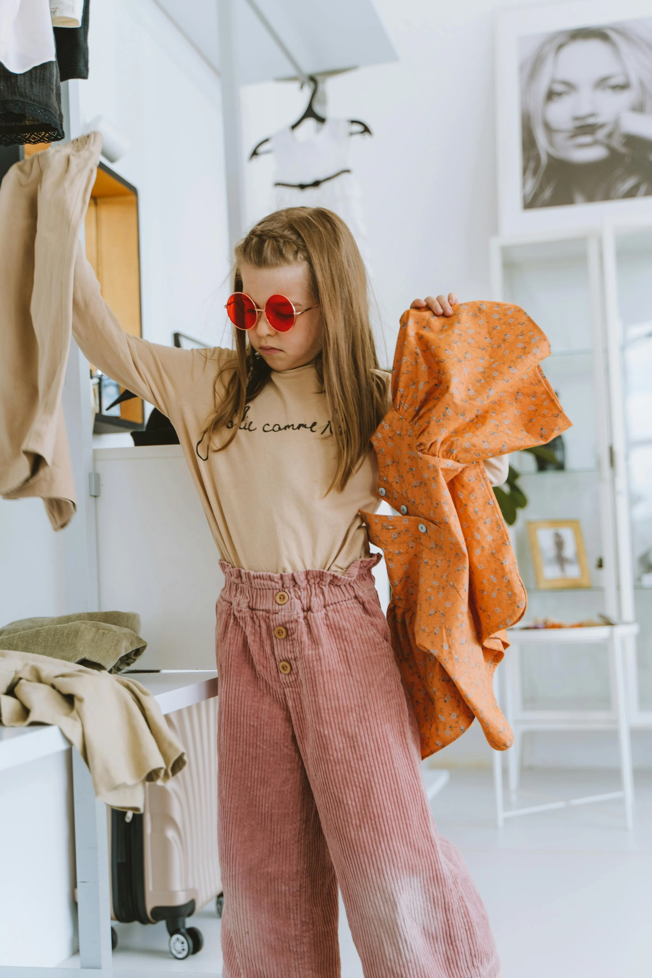 A young girl with long hair wearing red-tinted sunglasses, beige sweatshirt, and pink corduroy pants, shopping for clothes and holding a floral orange dress in a boutique or clothing store.