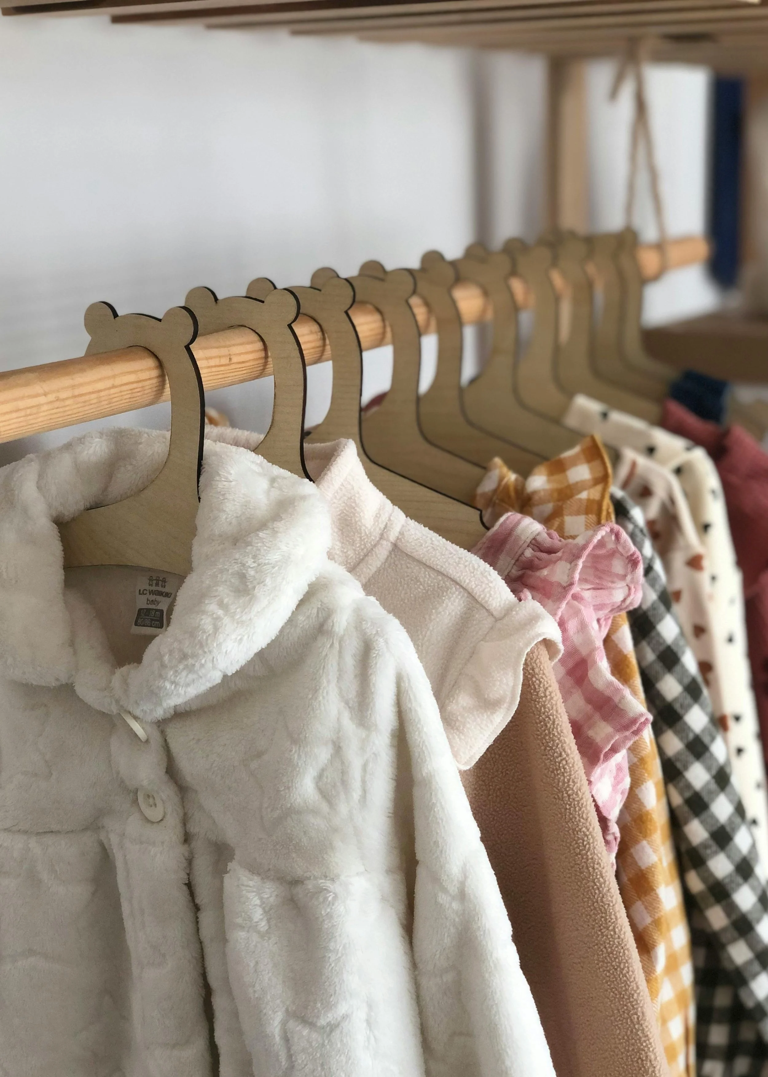 Kids' clothing on hangers, including plush white jackets, pink and tan dresses, and patterned shirts, hanging on a wooden rod in a retail store.