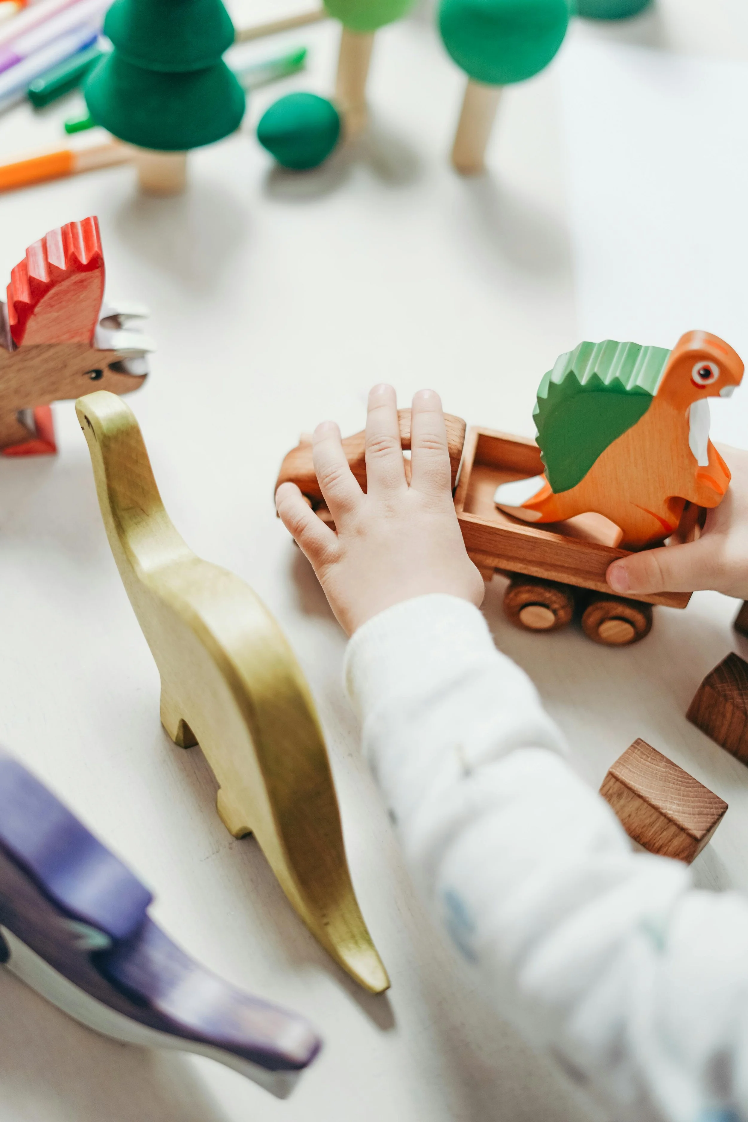 Close-up of a child's hand reaching toward toy wooden animals, including a green and orange dinosaur, on a white surface with other toys and colored markers in the background.