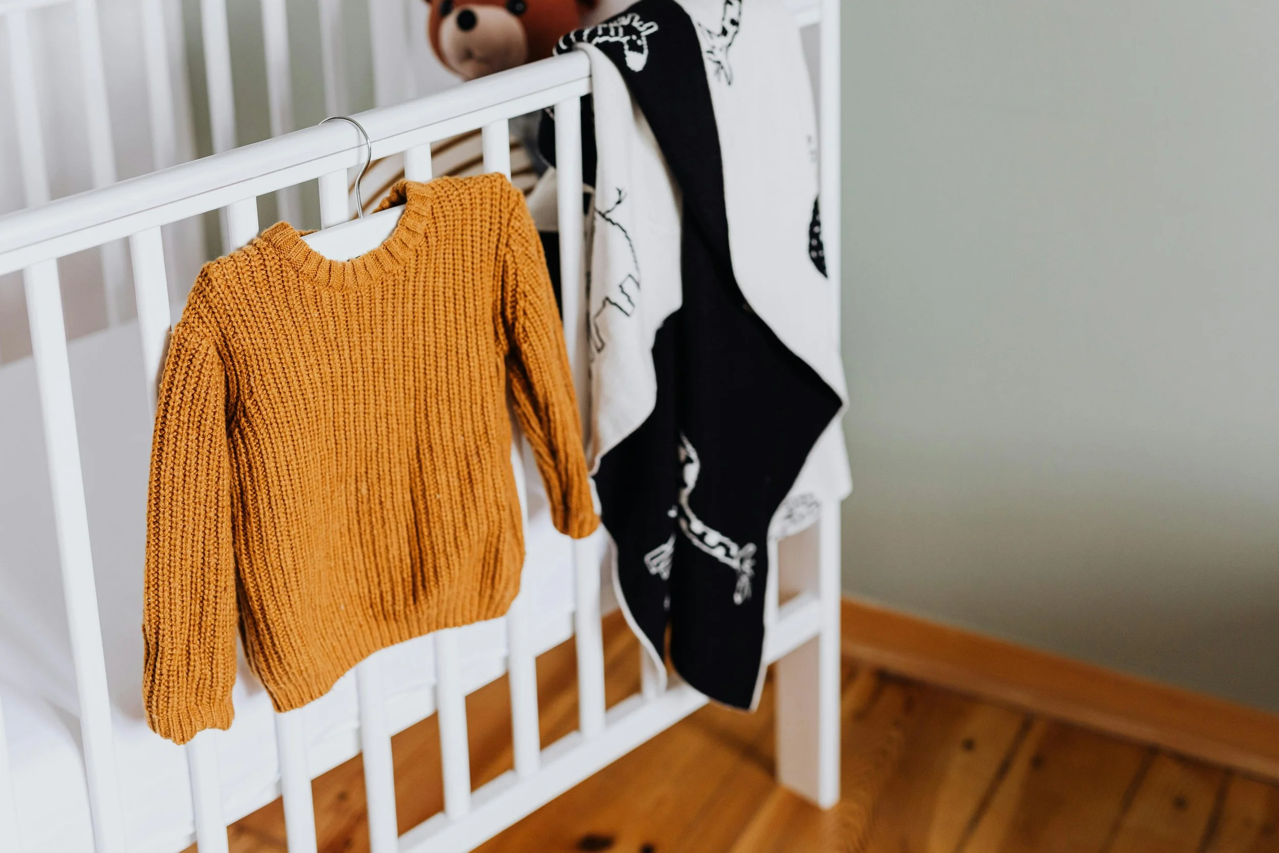 A yellow knitted sweater hanging on a white crib railing in front of a stuffed bear with a brown face and black eyes, and other clothing items hanging on the crib.