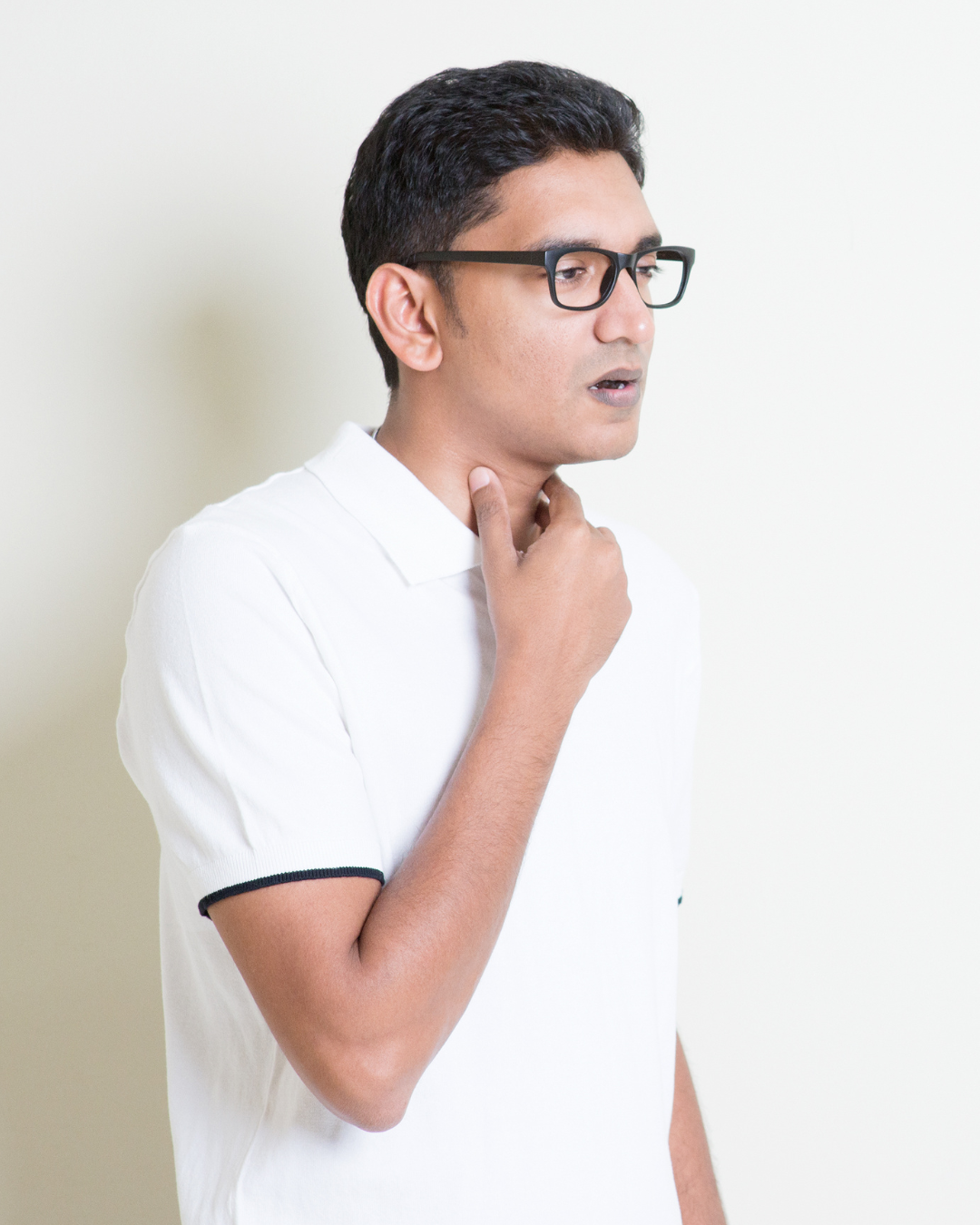 Man with black hair and glasses touching his throat, looks concerned, wearing a white polo shirt, standing against a white wall.