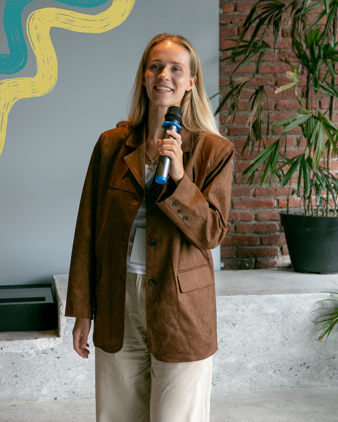 A woman with long blonde hair holding a microphone, smiling, dressed in a brown blazer and light-colored pants, standing indoors with a brick wall and potted plant in the background.