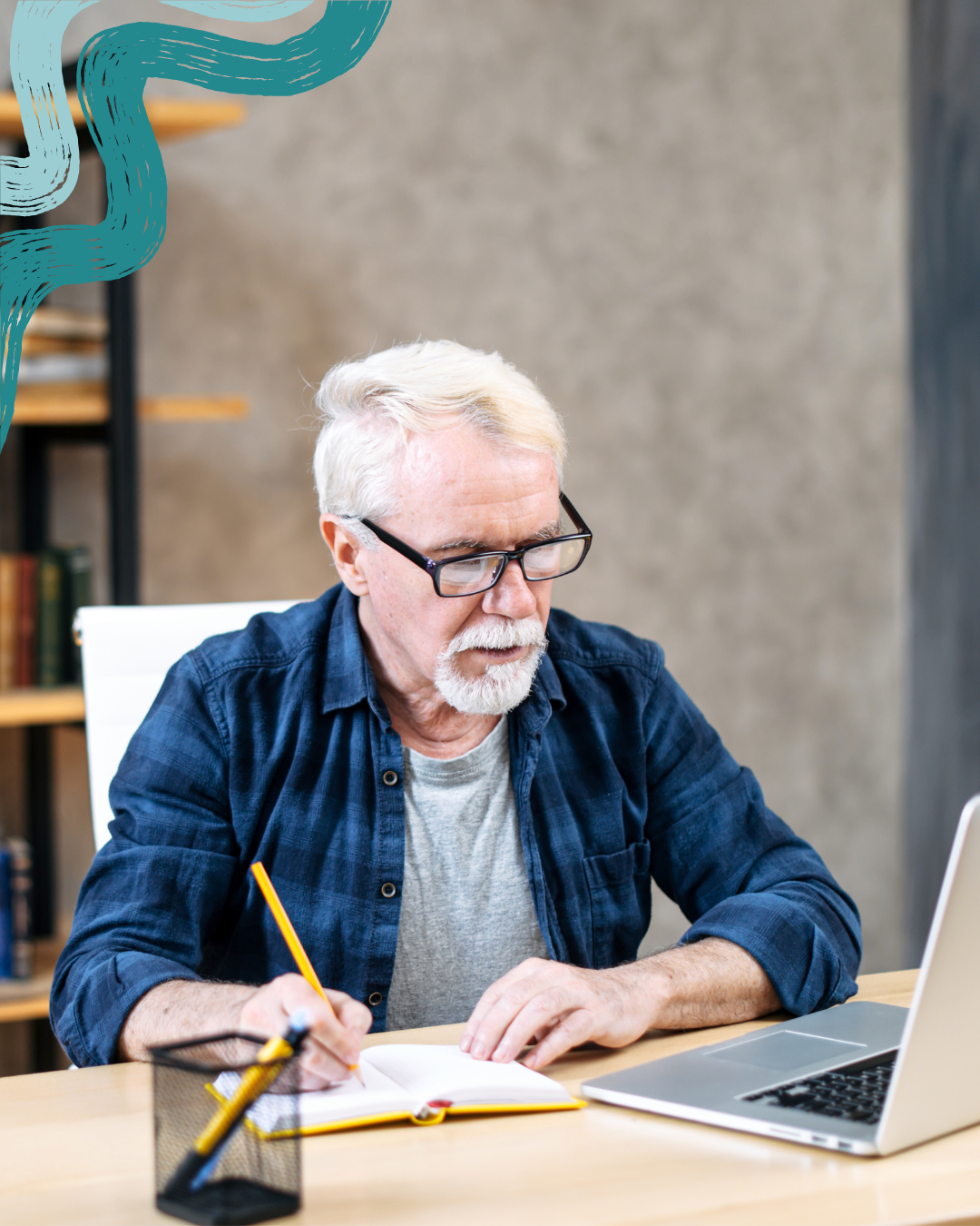 An elderly man with white hair, glasses, and a beard, wearing a gray t-shirt and blue plaid shirt, sitting at a desk with a laptop, notebook, and pens, writing with a yellow pencil.