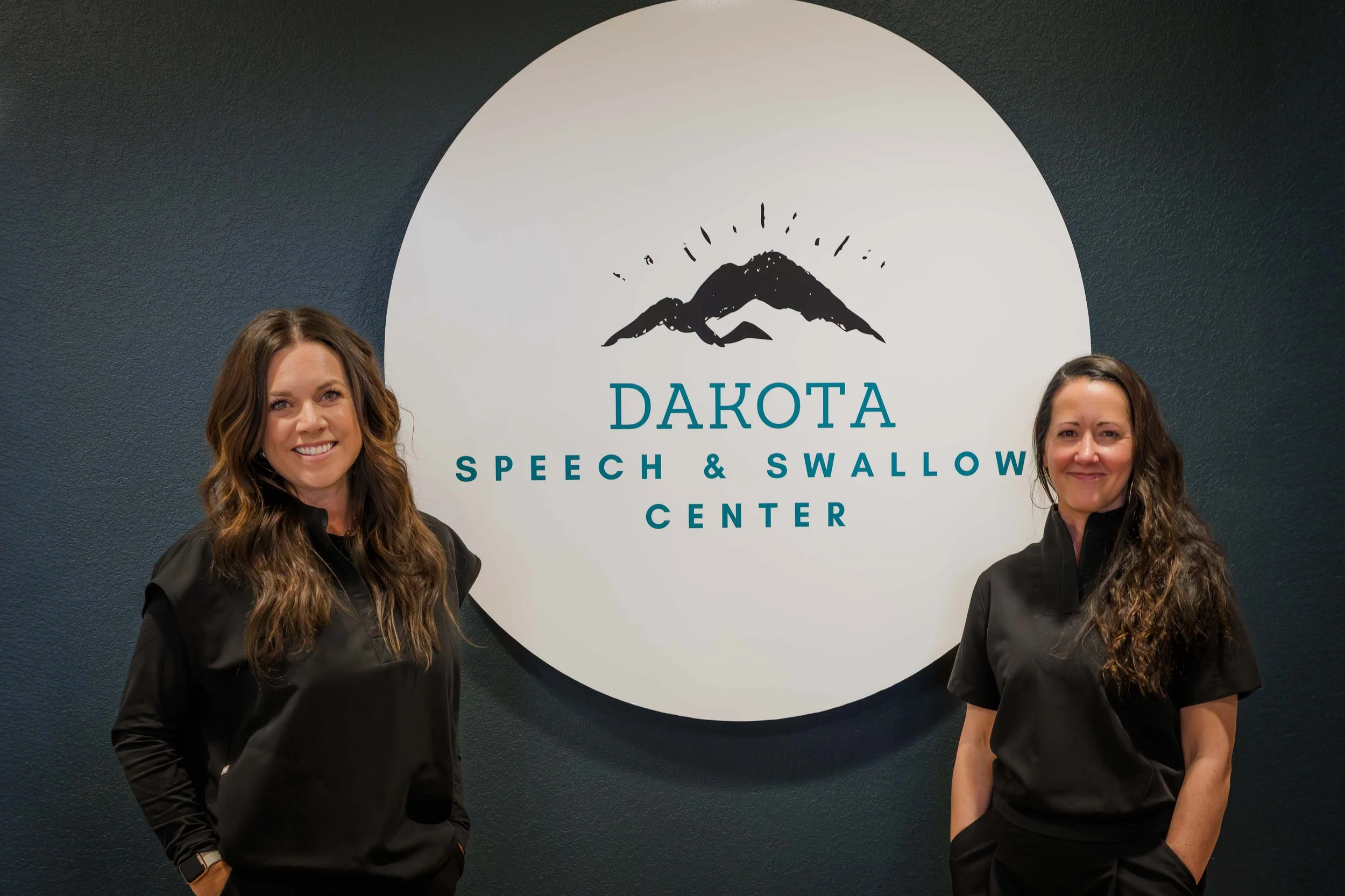 Two women standing in front of a sign for the Dakota Speech & Swallow Center, smiling at the camera, with dark hair and wearing black jackets.