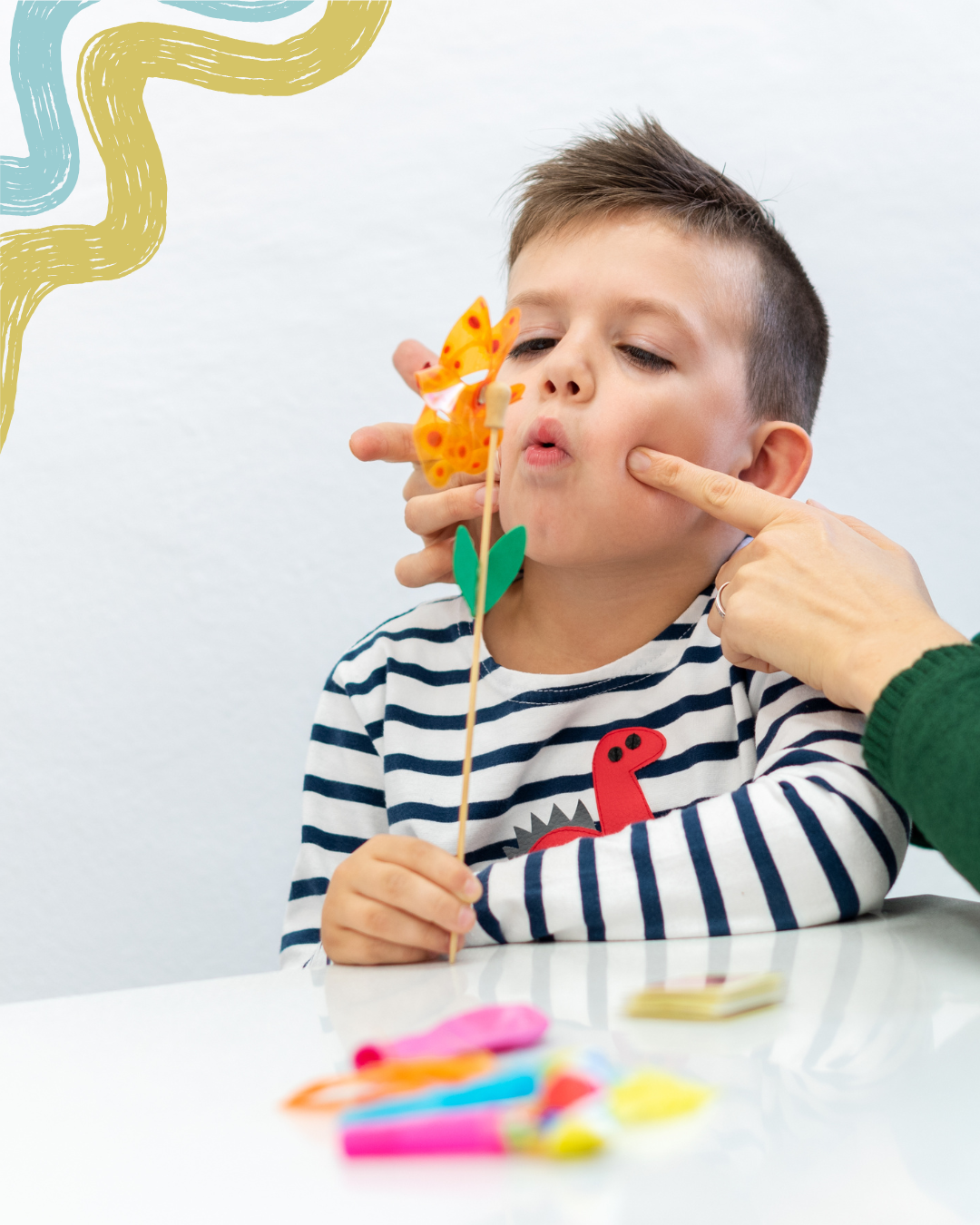 A young boy with short brown hair and wearing a striped shirt is holding a butterfly craft puppet made of colorful foam and sticks. An adult is gently touching his cheek while he looks at the puppet. There are various foam pieces on the white table in front of them.