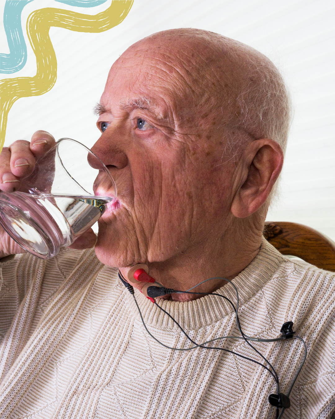 An elderly man with a bald head is drinking water from a glass, wearing a beige sweater and headphones with wires around his neck.