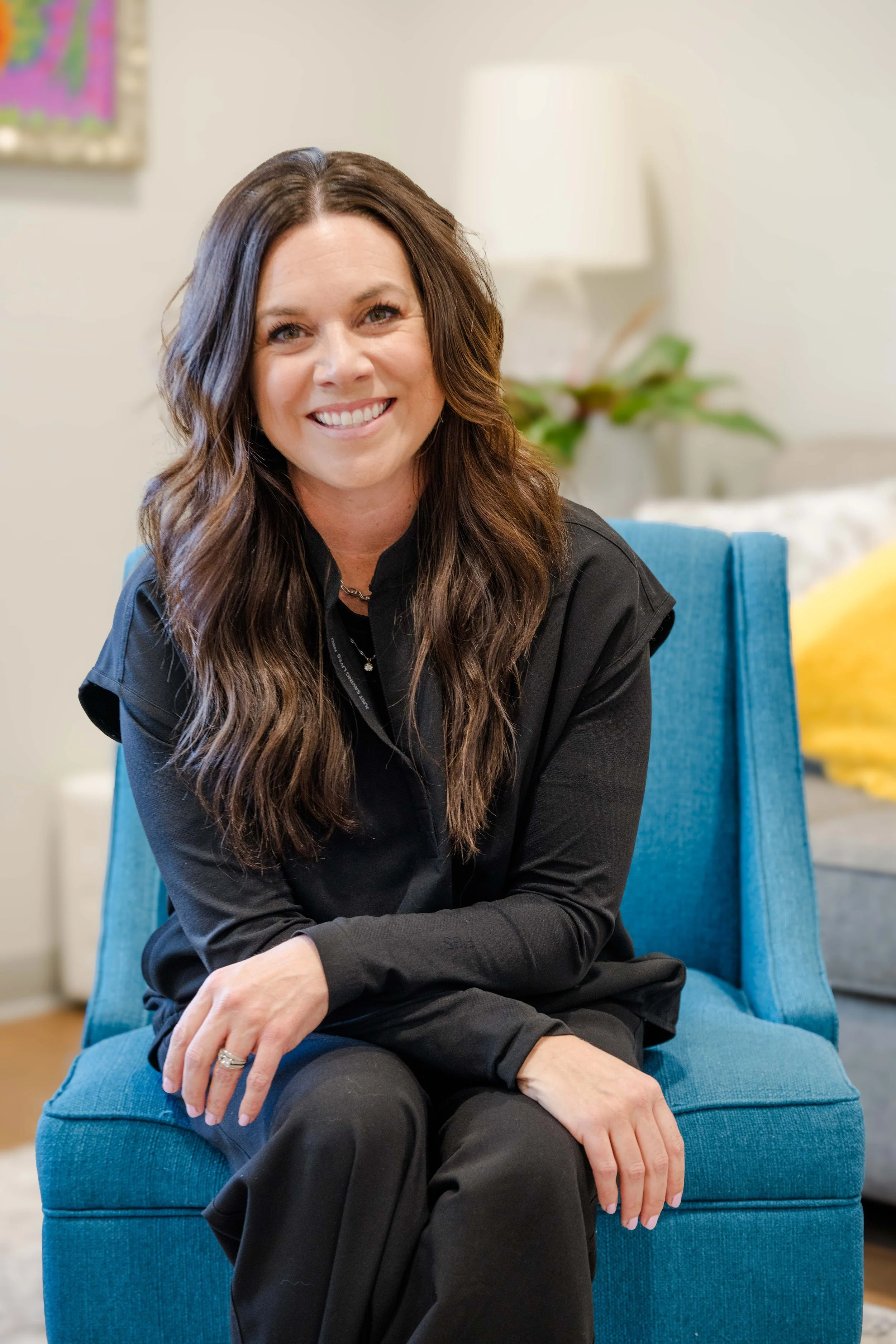 A woman with long, wavy brown hair, wearing a black long-sleeved shirt, sitting on a blue chair, smiling, in a cozy room with a lamp, plant, and a colorful wall hanging in the background.