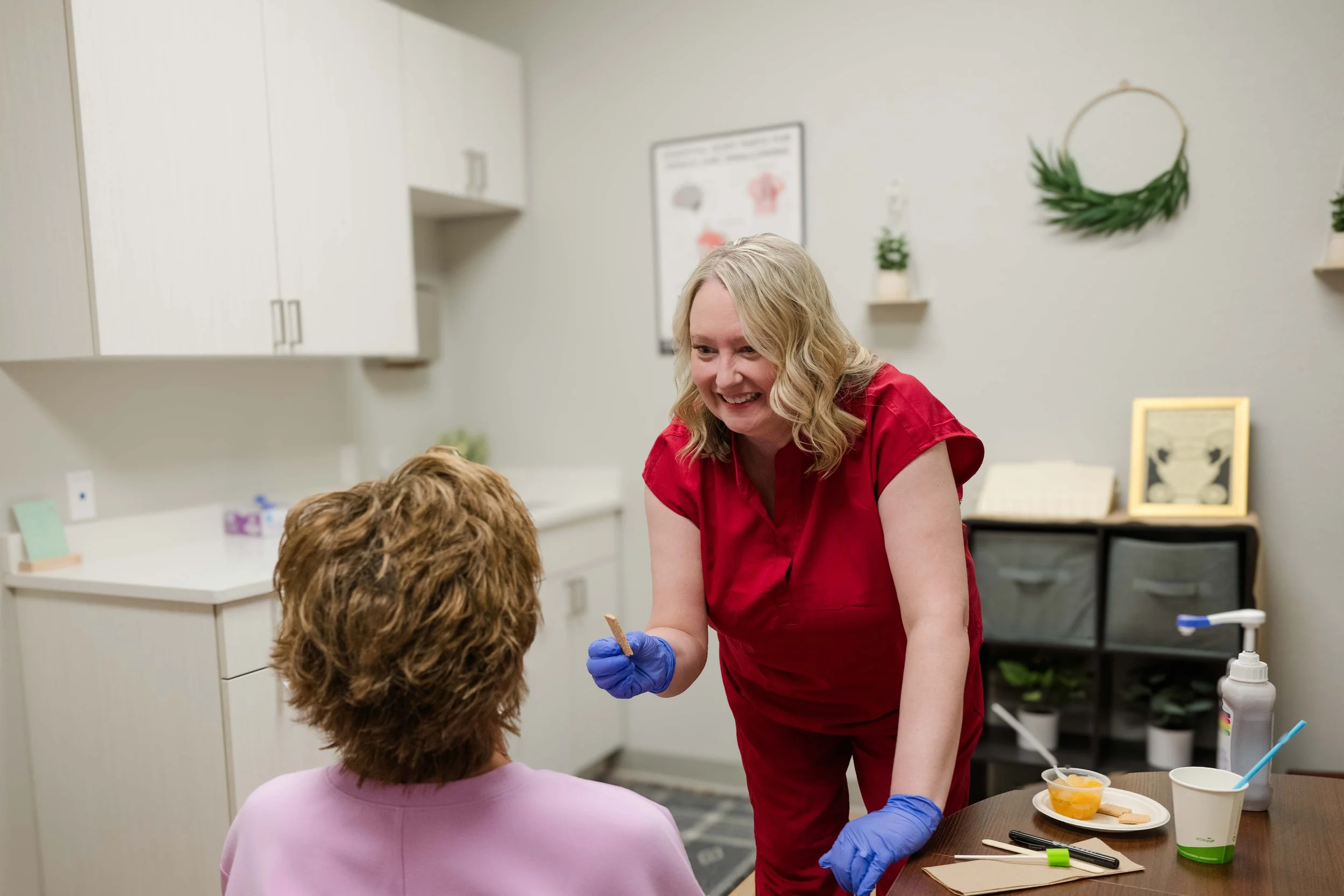 A woman with blonde hair wearing a red shirt and blue gloves, smiling and engaging with a young child with curly hair, in a medical or dental office. The woman is holding a small device and the child is sitting at a table with various items, including cups, a plate with snacks, and stationery.