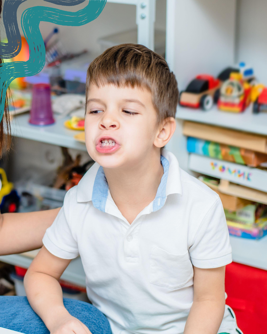 A young boy making a face or grimace in a playroom with toys in the background.