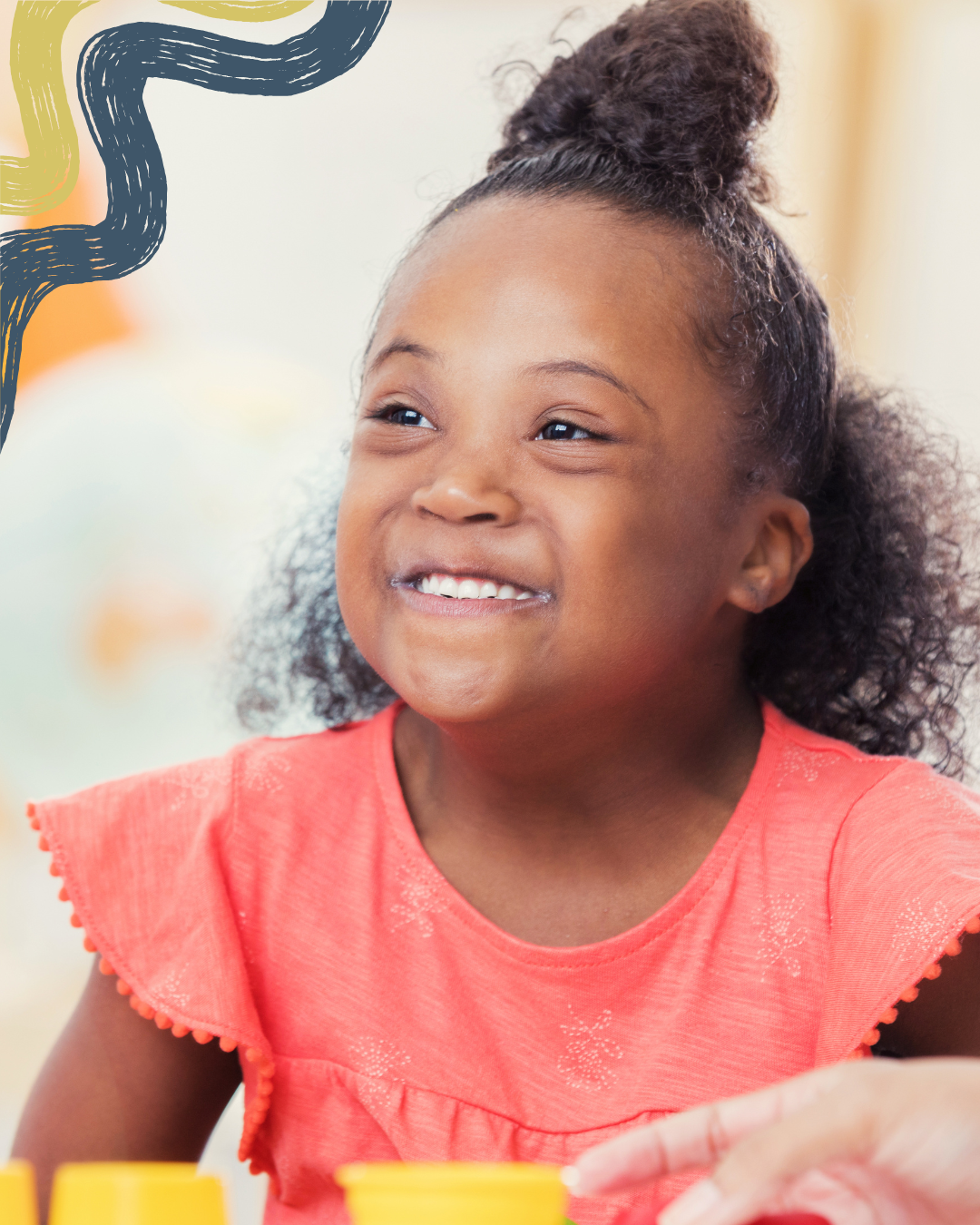 A young girl smiling and sitting at a table with yellow containers in front of her, wearing a coral pink top.