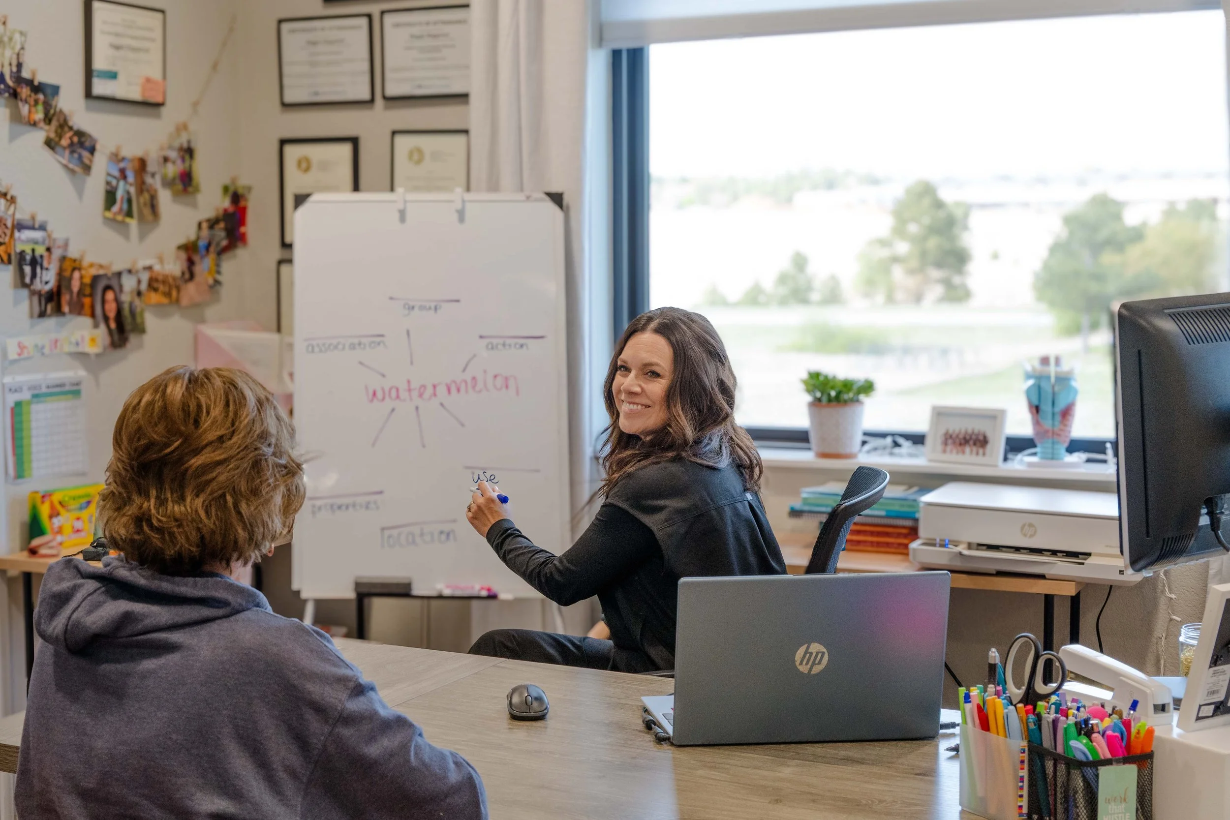 A woman is sitting at a desk, smiling, while a person with curly hair faces her. Behind the woman, a whiteboard with notes about watermelons is visible. The room has a window, plants, framed certificates, and decorative pictures on the wall.