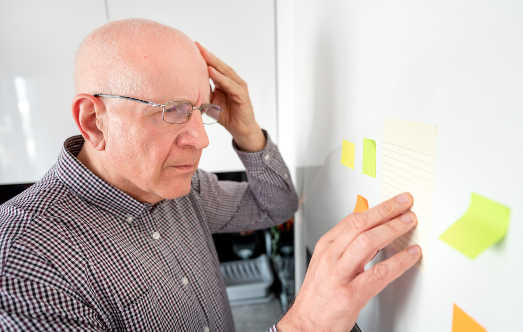 An older man with glasses and a checkered shirt looks stressed while placing sticky notes on a whiteboard in an office.