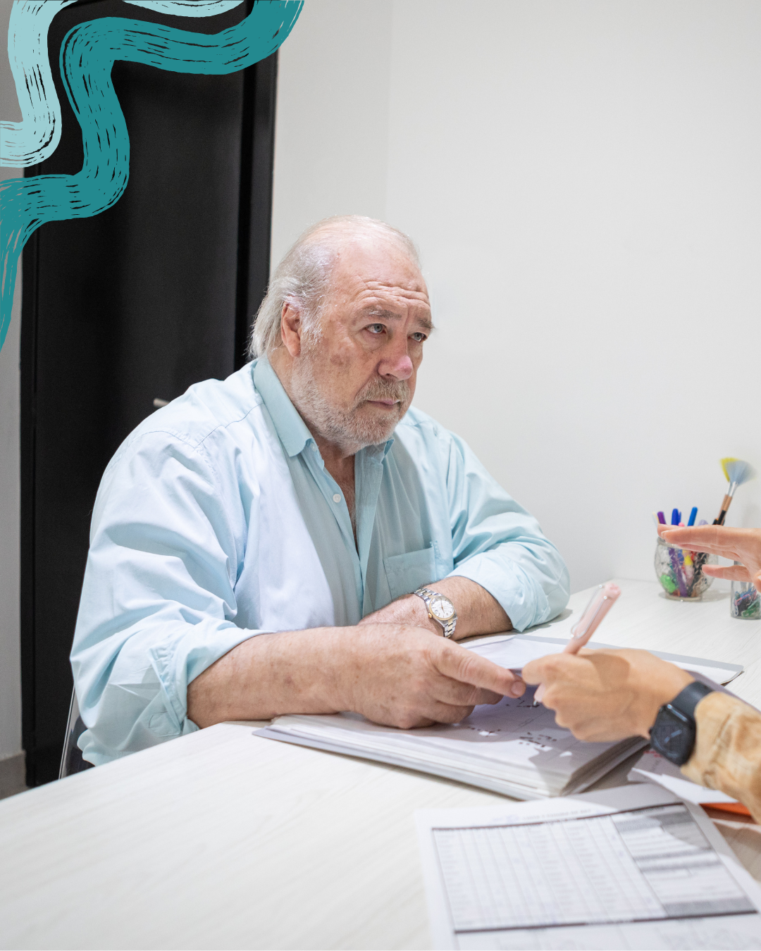 An elderly man with gray hair and a beard, wearing a light blue shirt, sitting at a table and looking at a document held by a person with a smartwatch. The table has papers, and there are colorful pens in a cup on the side.