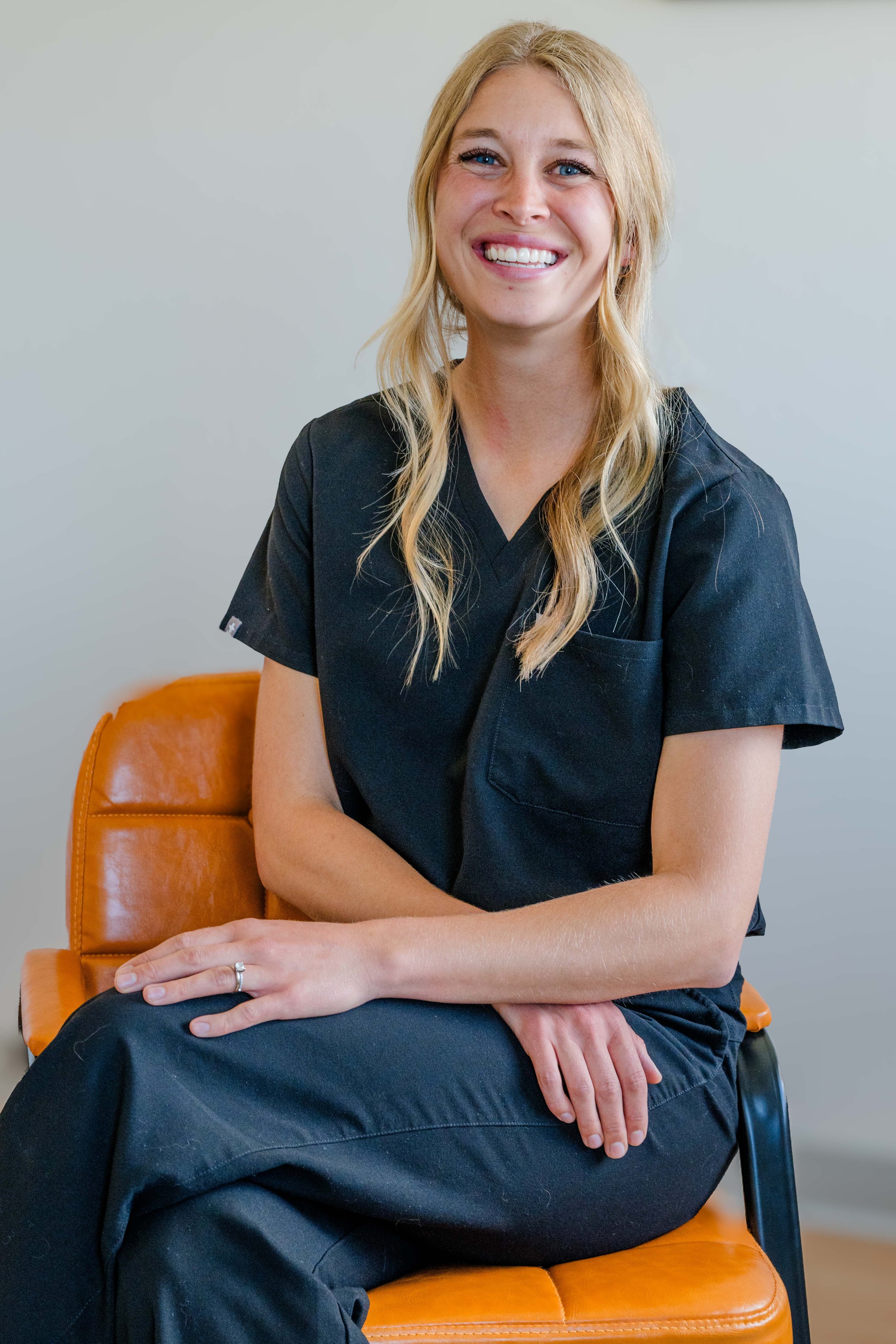 A smiling woman with blonde hair wearing a black medical scrubs, sitting on an orange chair.