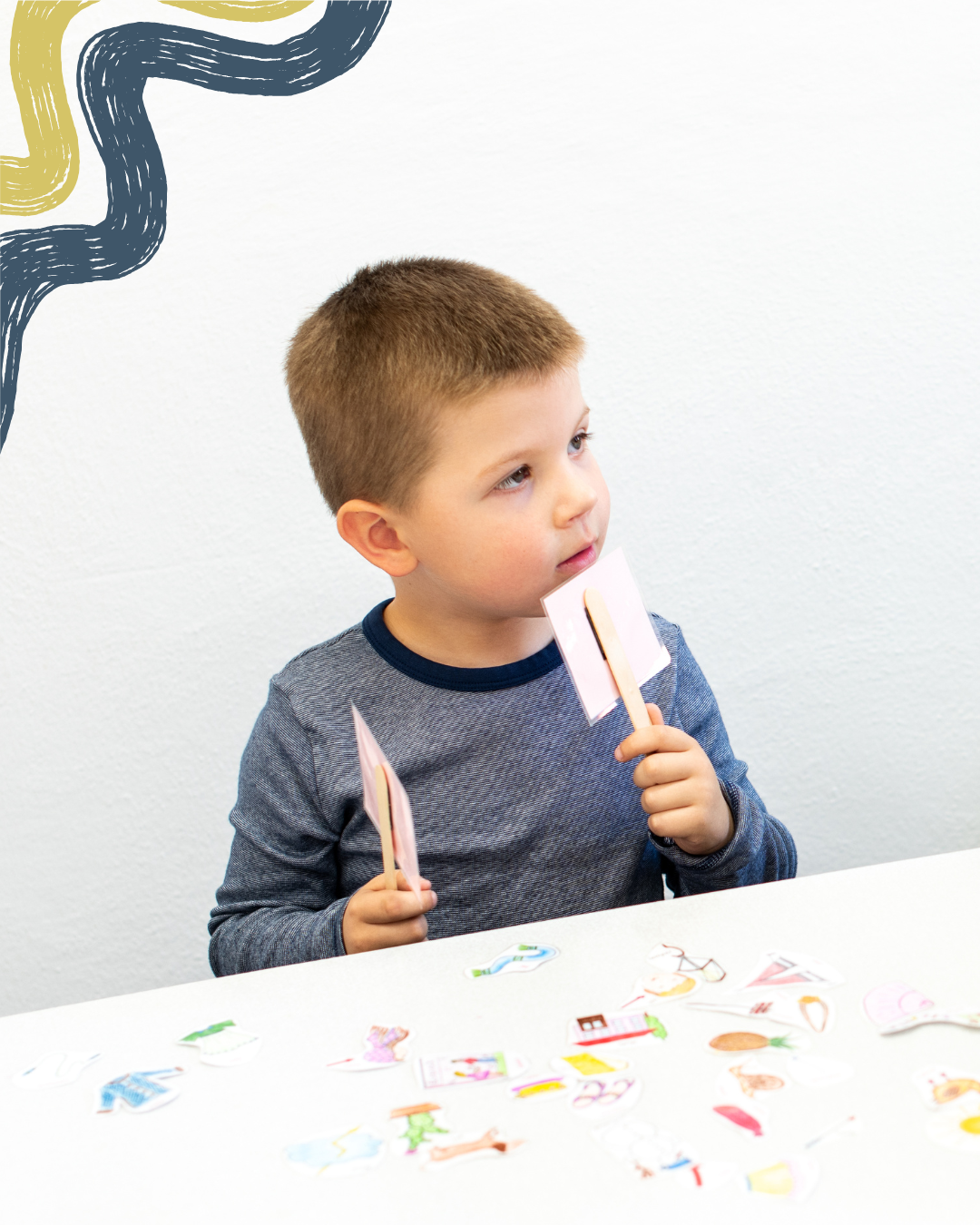 A young boy with short brown hair holding two paper cards on sticks, sitting at a table with colorful paper cutouts, in front of a white wall with abstract black and gold artwork.