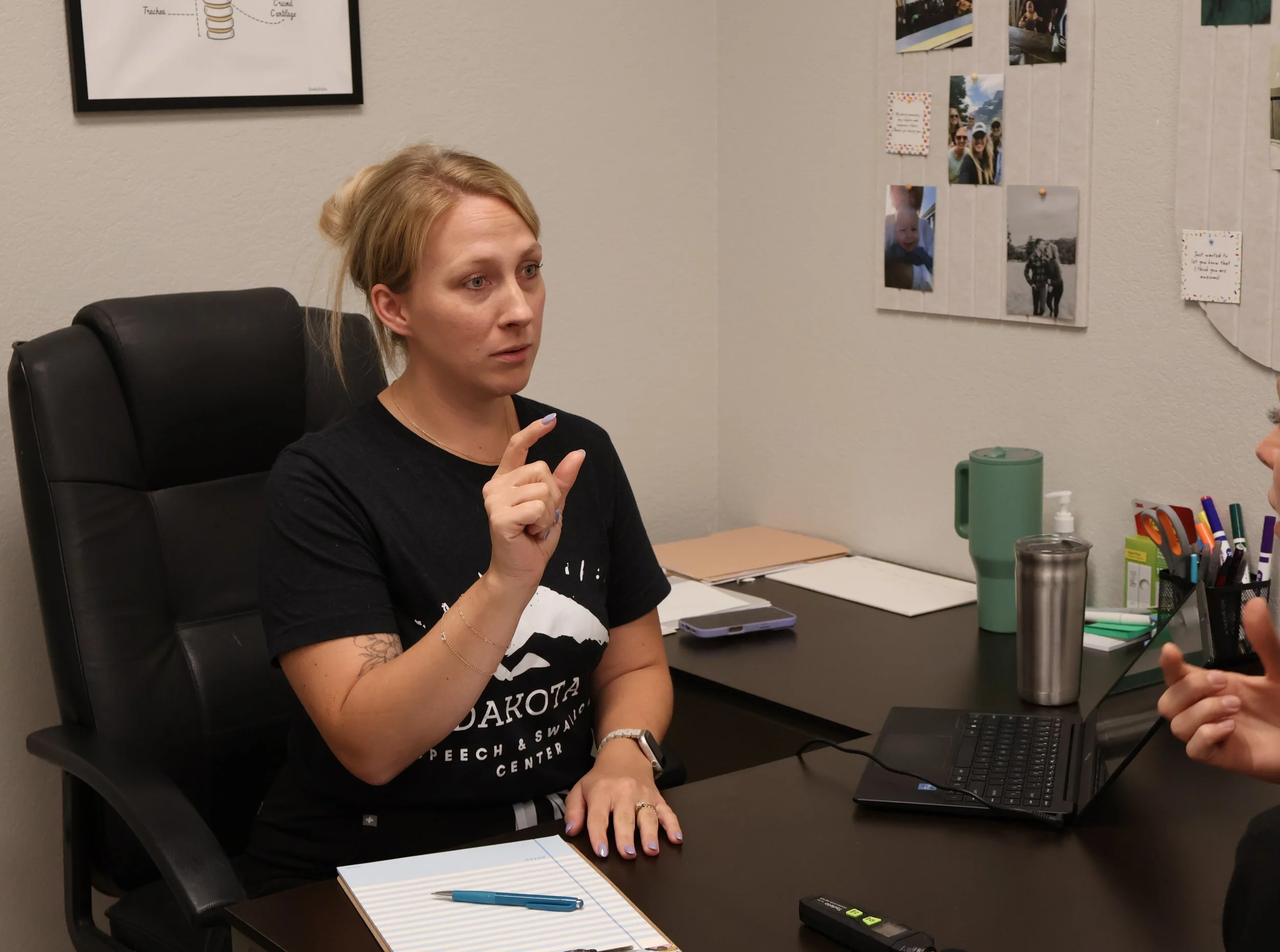 A woman with blonde hair sits at a desk engaged in conversation, with a notepad, pen, and a laptop in front of her. Photographs are pinned on the wall behind her, and various office supplies are on the desk.