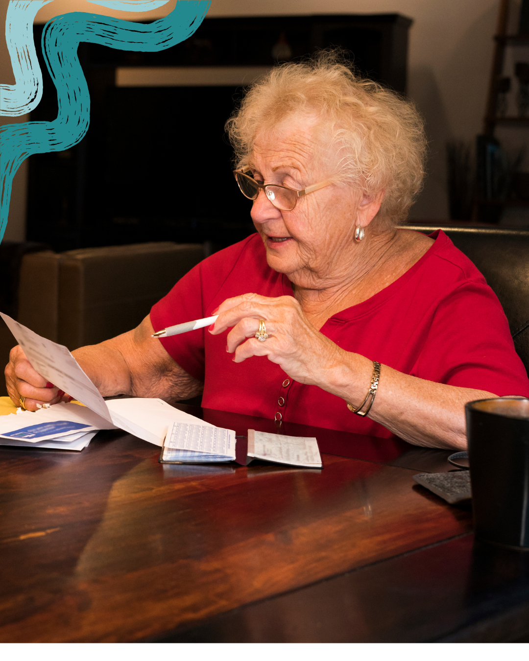 An elderly woman with curly blonde hair, glasses, and hoop earrings sitting at a wooden table, looking at documents with a pen in her hand. She is wearing a red shirt, a bracelet, and a ring, and appears to be reviewing paperwork or bills.