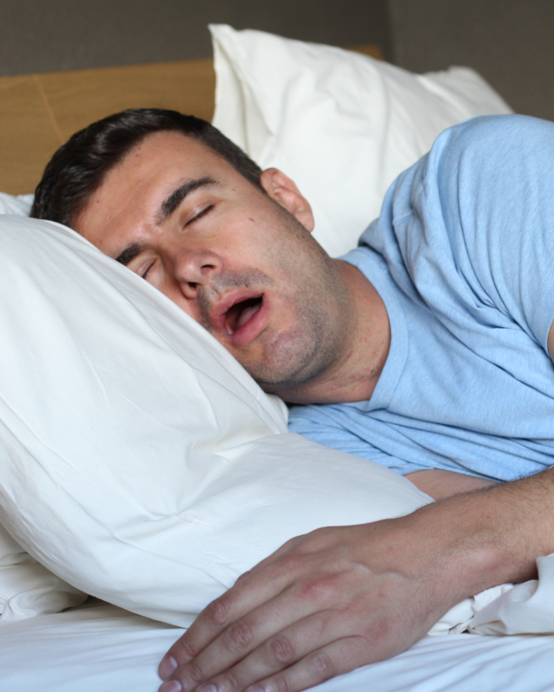 Man sleeping on a bed, lying on his side with his head on a white pillow, wearing a light blue shirt.