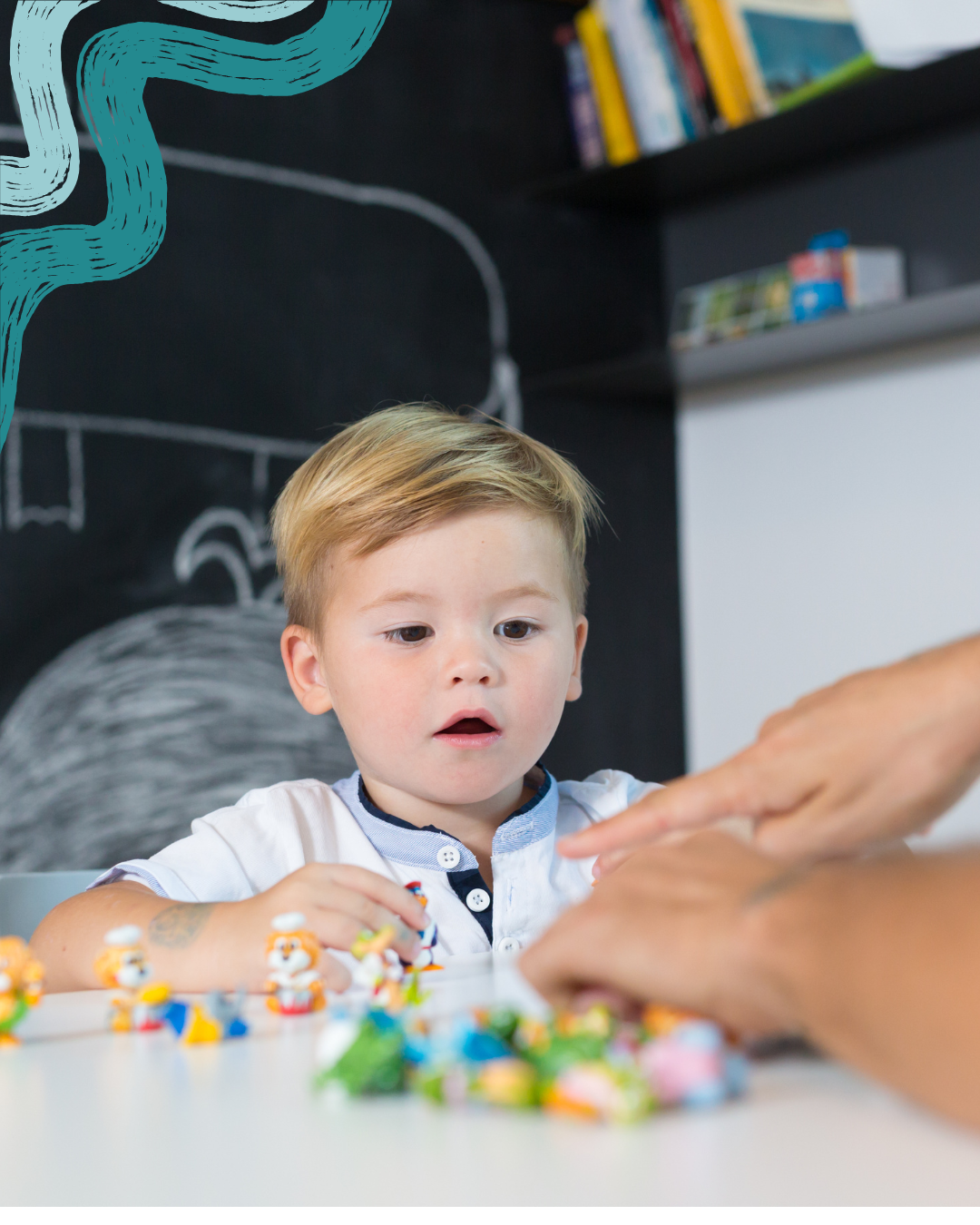 A young boy with blond hair sits at a white table, playing with colorful figurines and toys while an adult's hand reaches towards his toys. In the background, there is a black wall with chalk drawings and a shelf with books and boxes.