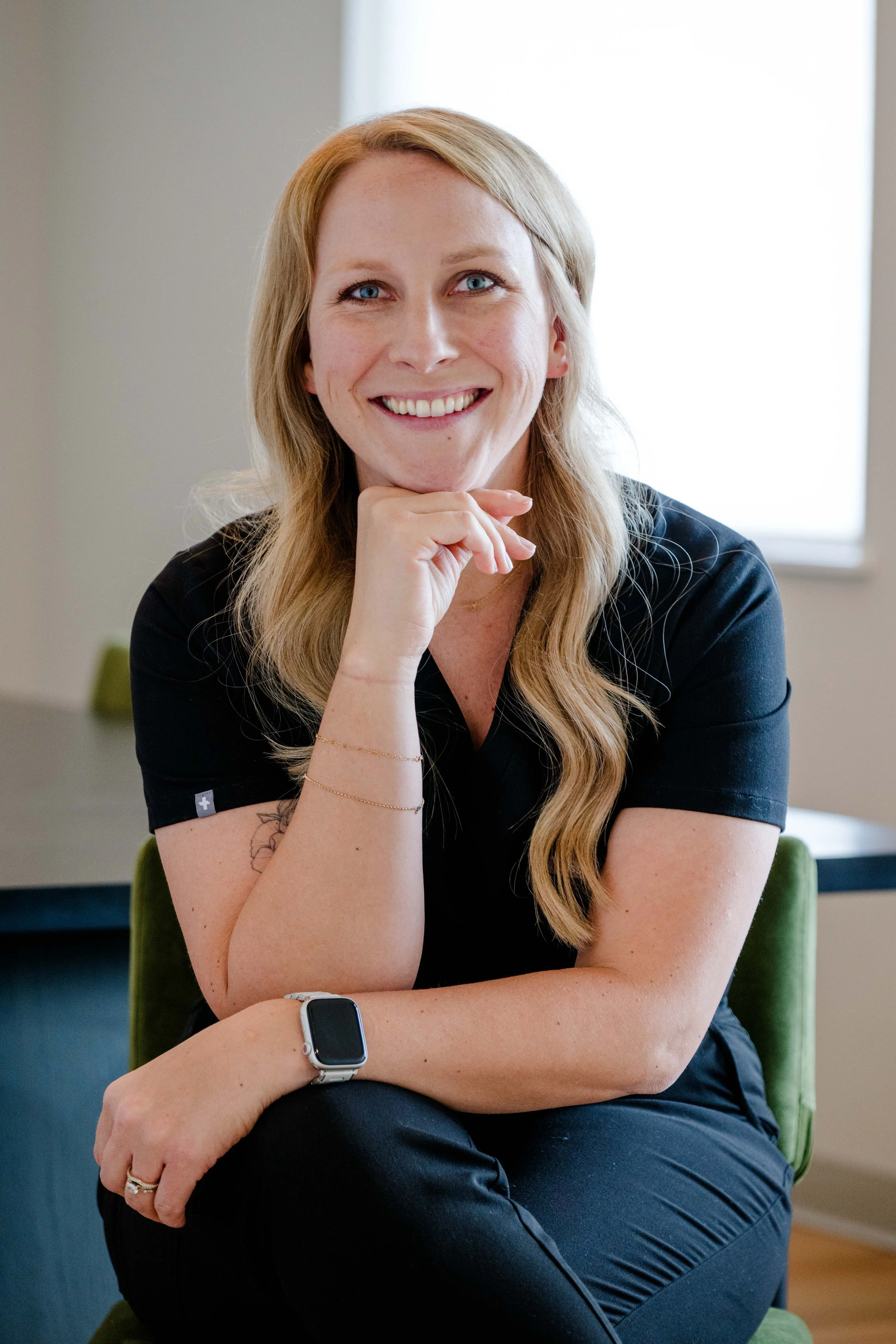 A woman with long blonde hair, blue eyes, and a bright smile, sitting on a green chair indoors. She is wearing a black shirt and has a smartwatch and rings on her right hand, which is resting on her chin.