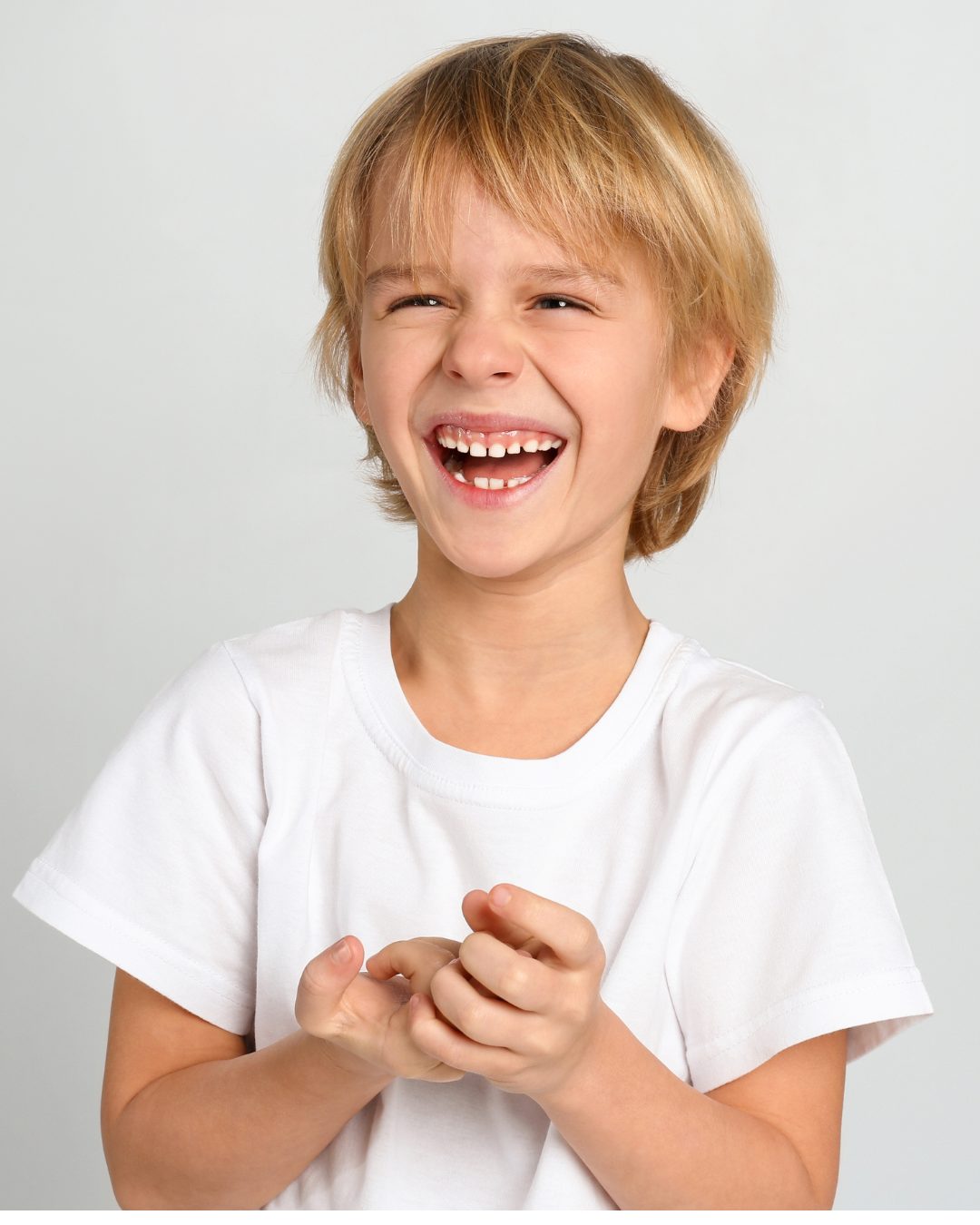 A young boy with blond hair, smiling and showing missing front teeth, wearing a white t-shirt, against a gray background.