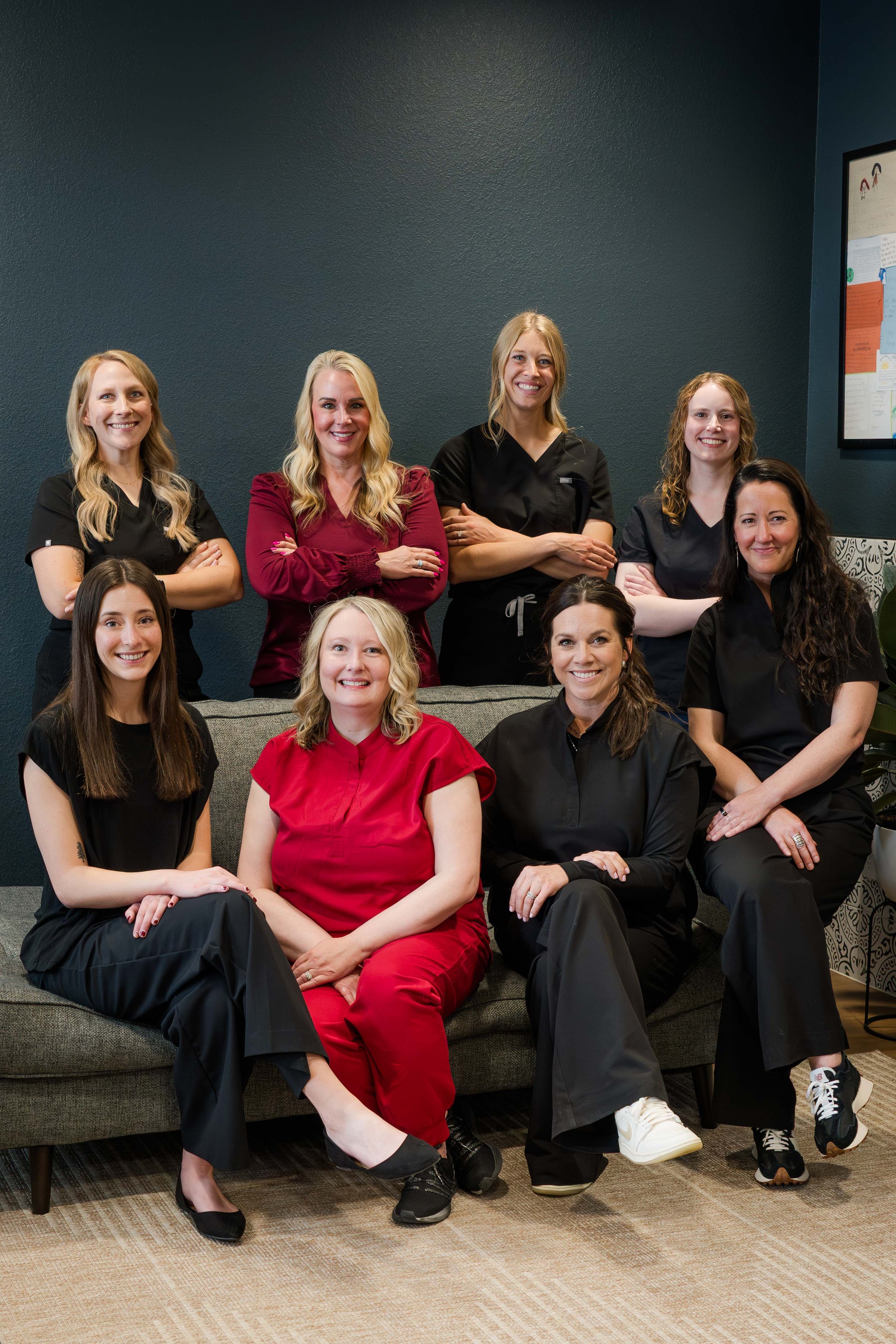 A group of ten women, most wearing black scrubs and one in red scrubs, sitting and standing in a room with dark teal walls, smiling at the camera.