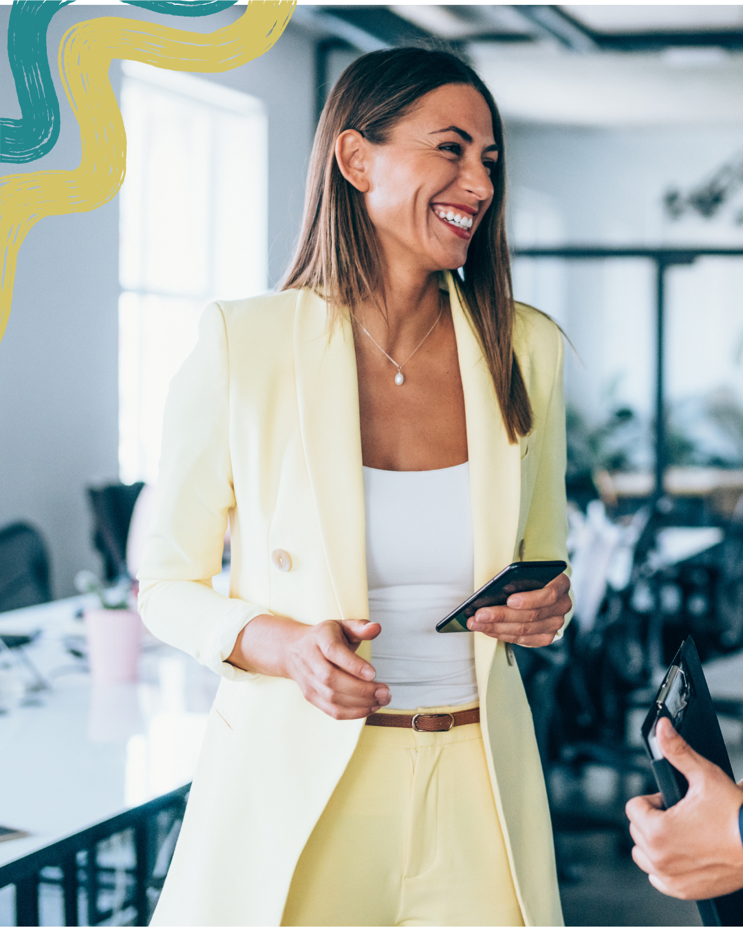 A woman in a yellow suit smiling and holding a smartphone in a bright, modern office.
