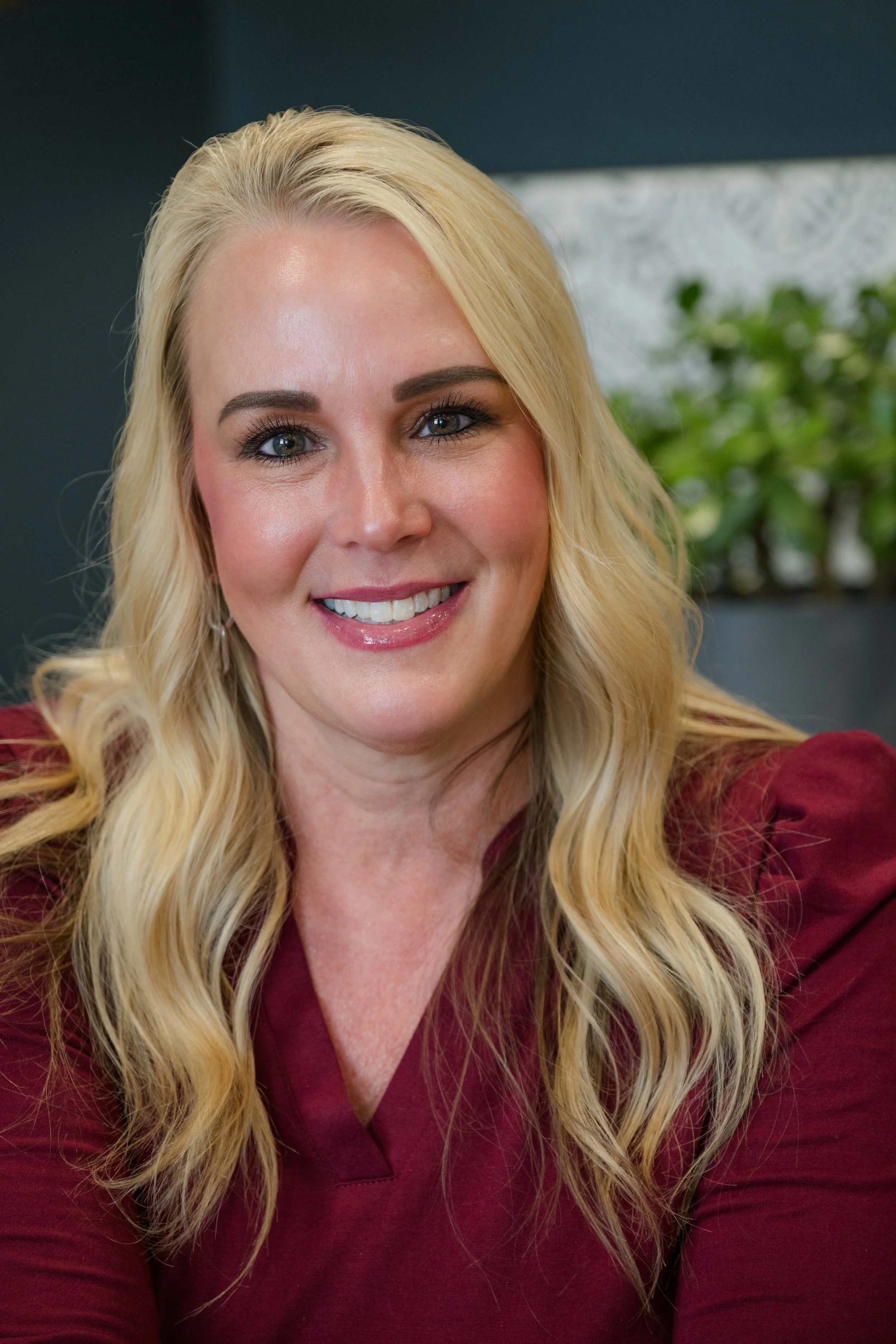 Close-up of a woman with long blonde hair, blue eyes, and a bright smile, wearing a maroon top, against a background with a green plant and a dark-colored wall.