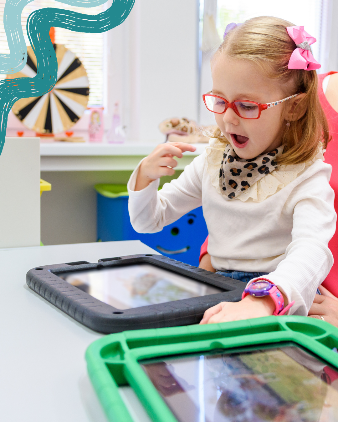 A young girl with red glasses, pink bows in her hair, and a leopard-print scarf, reacts with surprise or excitement while using a tablet at a white table in a colorful classroom. There are two tablets with green and black protective cases on the table.