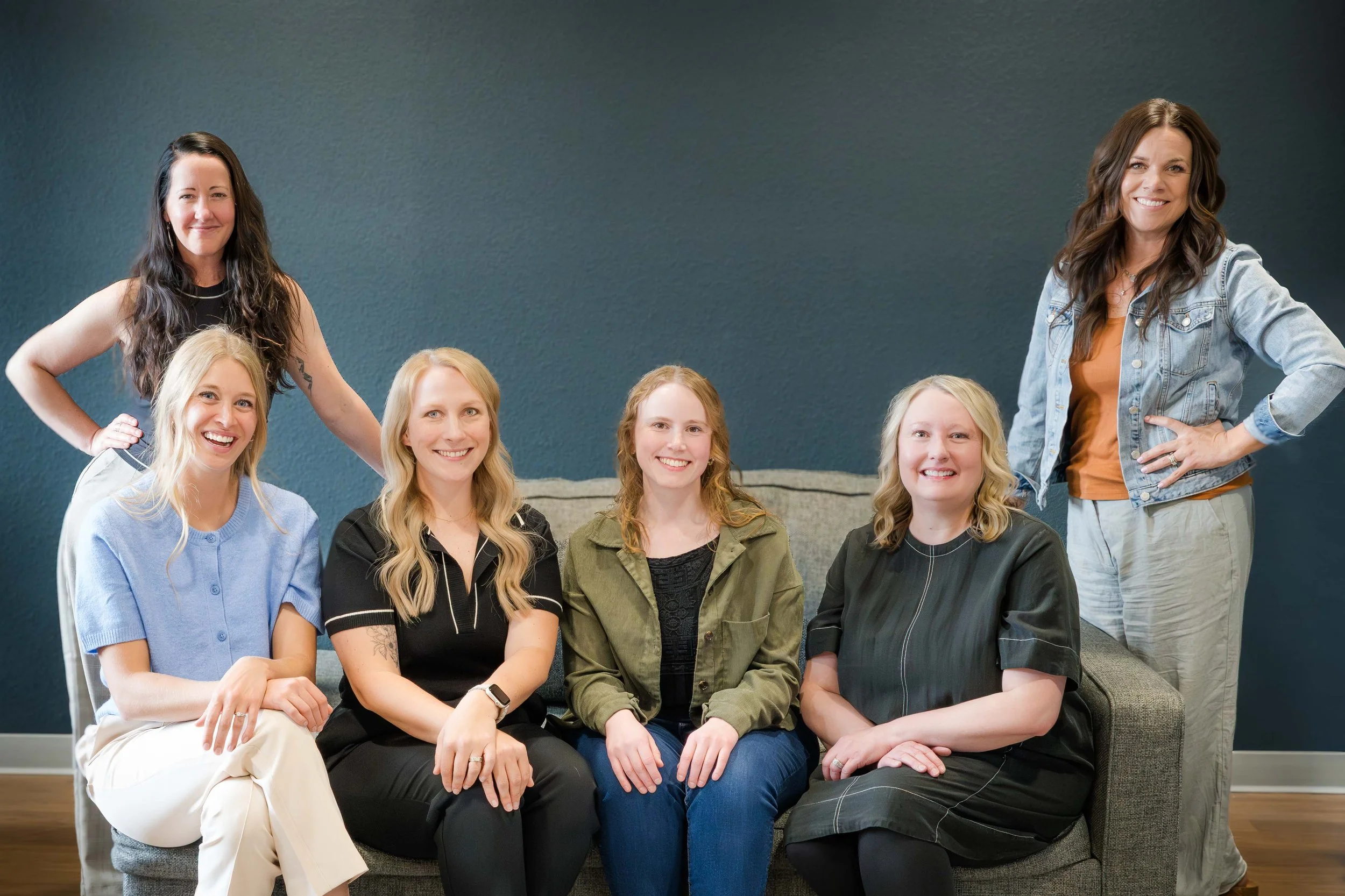 Group of seven women with long hair, sitting and standing on a couch in a living room, smiling at the camera, against a dark wall background.