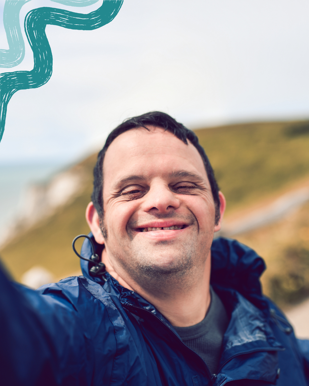 Man taking a selfie outdoors on a cloudy day with a blurred grassy hillside in the background, smiling with eyes closed, wearing a blue jacket.