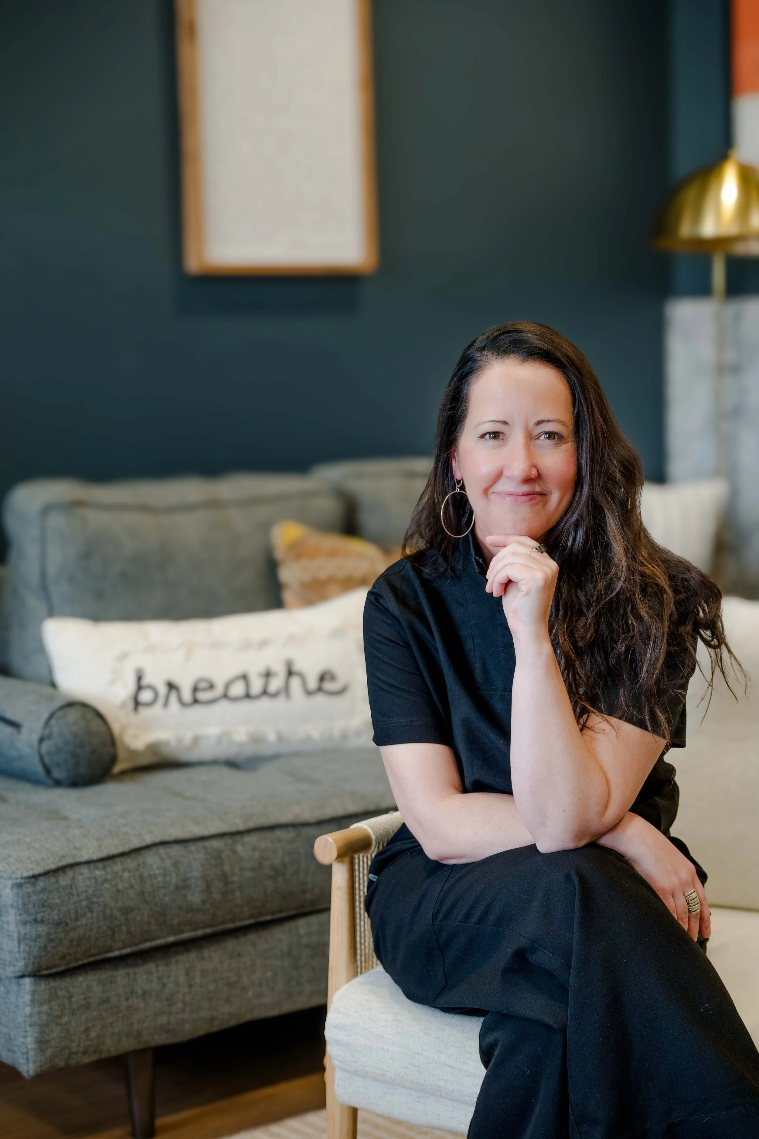 A woman with long brown hair, wearing hoop earrings and a black shirt, sitting on a small wooden chair in a cozy living room, smiling with her hand near her chin. In the background, there is a gray couch with a pillow that has the word 'breathe' written on it, and a dark green wall with a picture frame and a gold lamp.