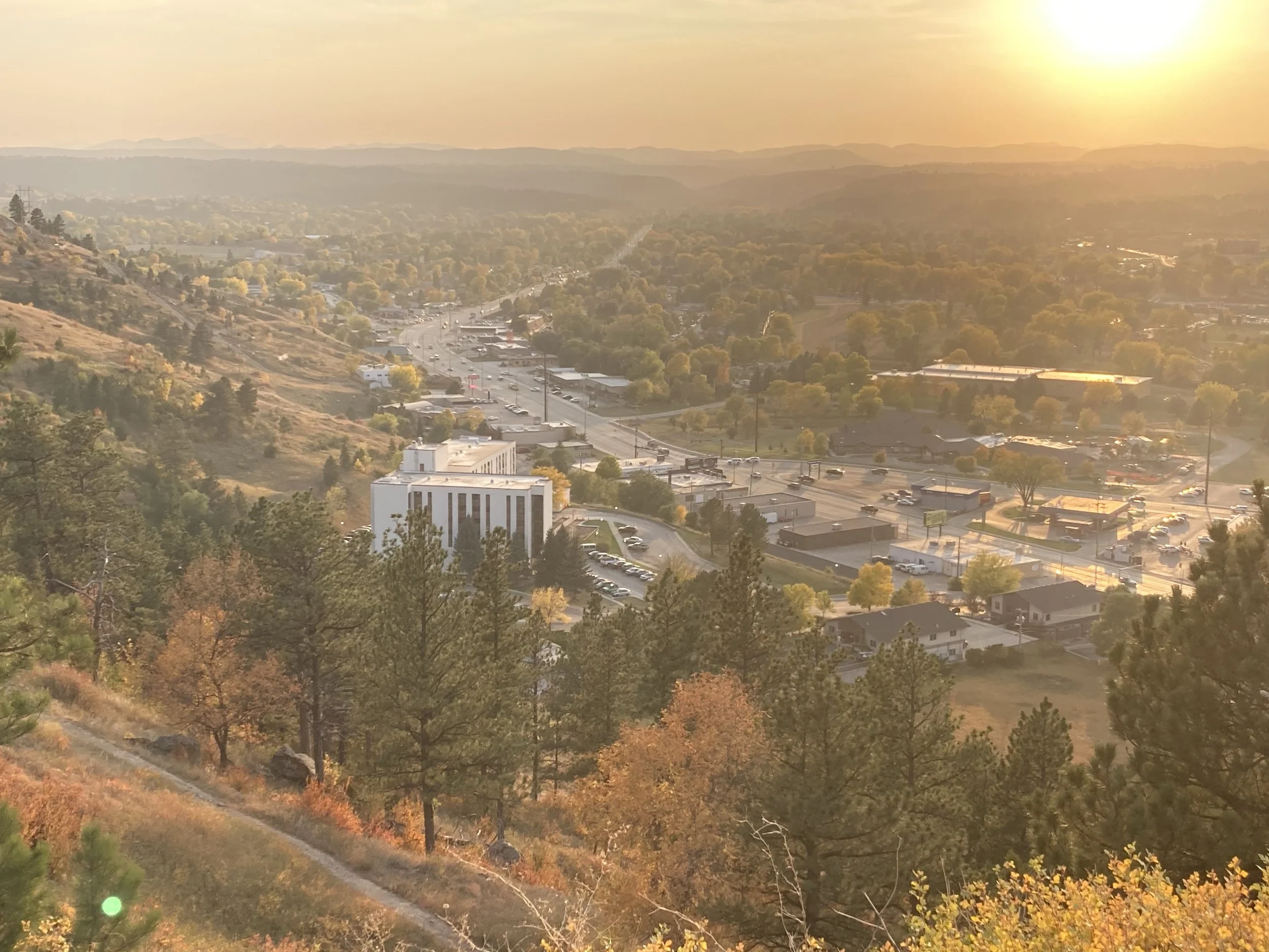 A scenic view of a hilly landscape at sunset with trees, roads, and buildings visible in the distance.
