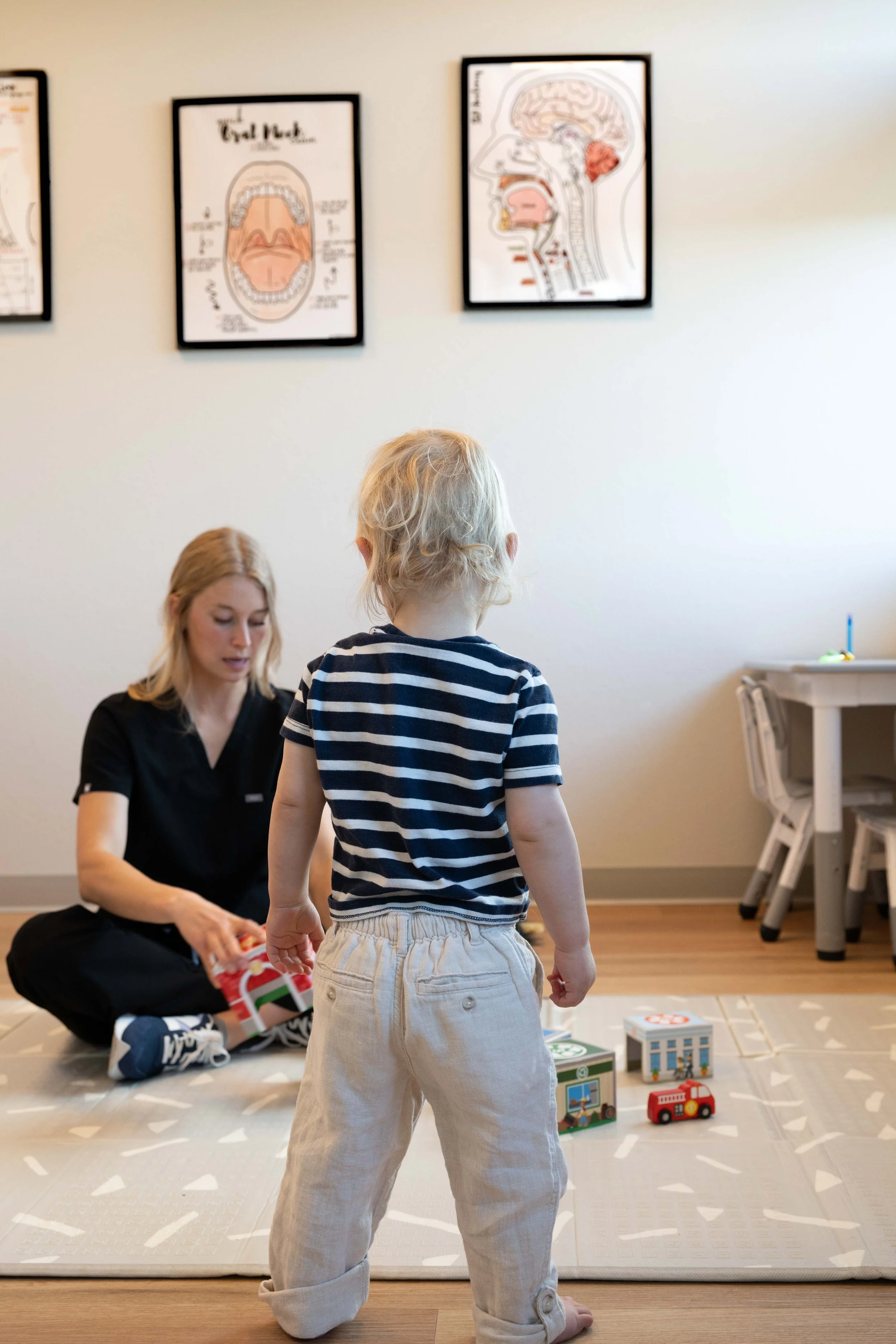 A young child with blonde hair, wearing a striped shirt and beige pants, stands on a play mat in a room, facing a woman who is sitting on the floor with toys and books nearby. Three framed anatomical diagrams hang on the wall behind them.