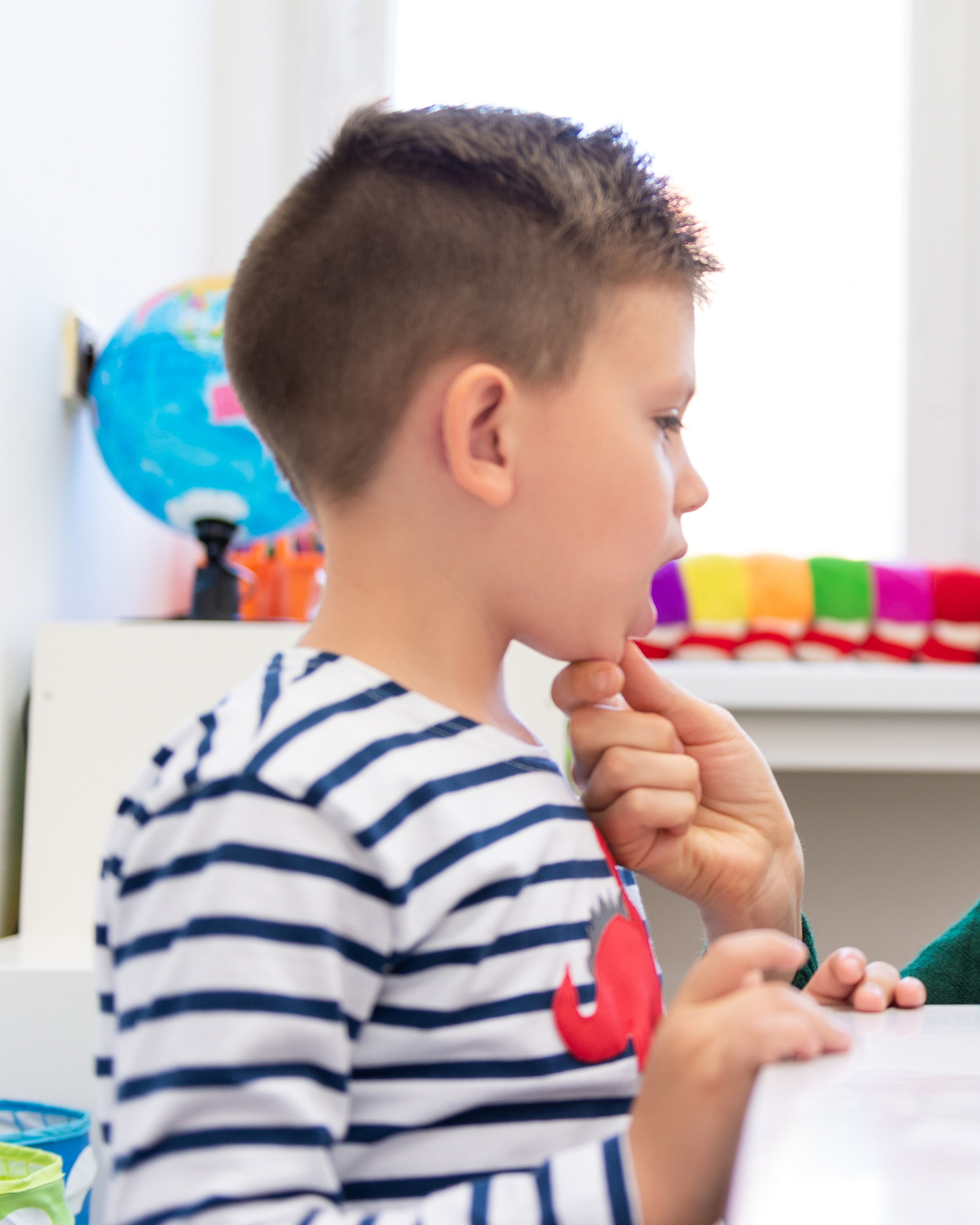 A young boy with short brown hair, wearing a striped shirt, is sitting at a desk with a thoughtful expression, resting his chin on his hand.