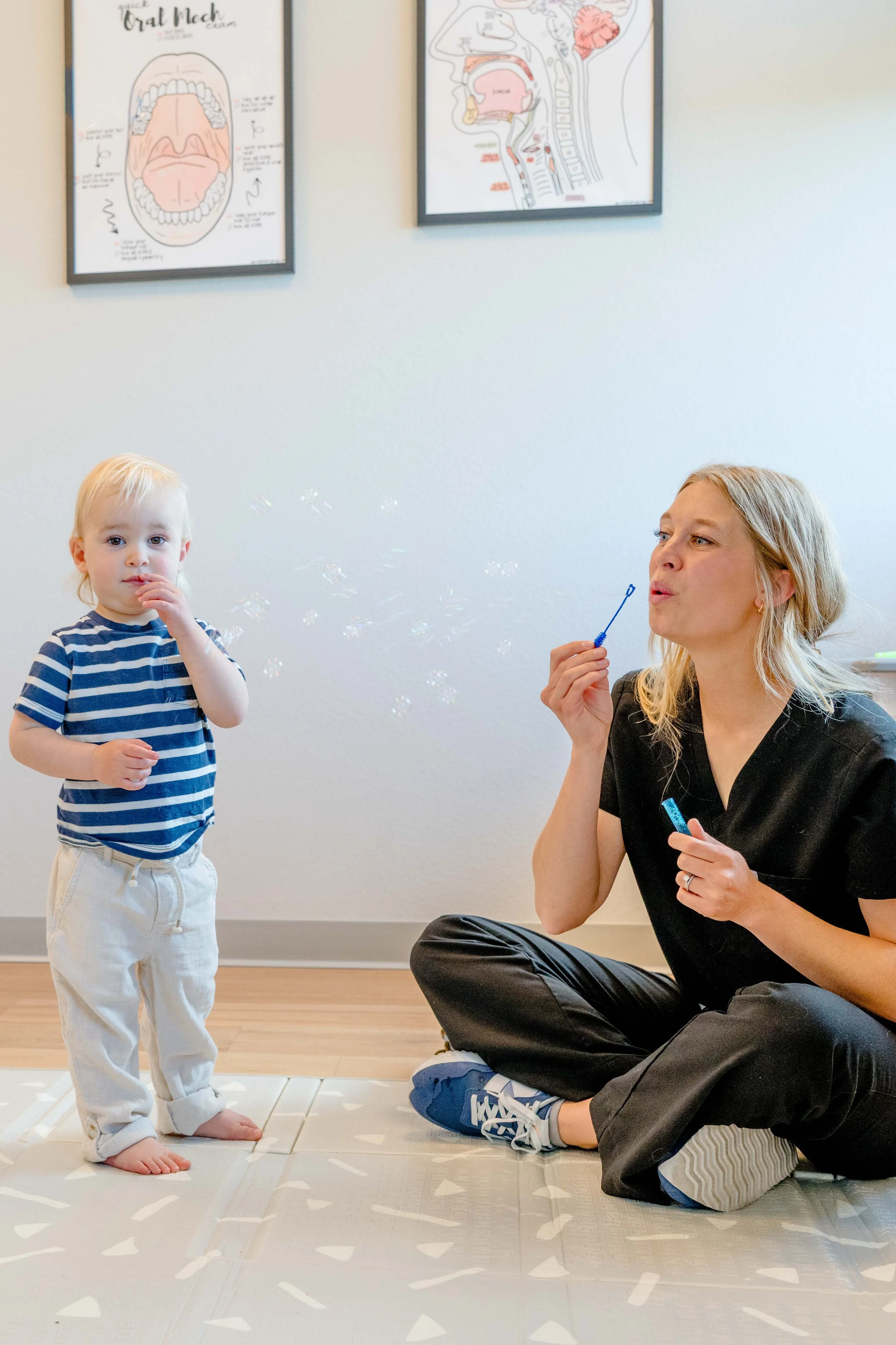 A young boy in striped shirt and white pants standing on a padded mat, with a woman sitting cross-legged on the floor blowing bubbles. The woman has blonde hair and is wearing black scrubs and blue sneakers. There are anatomical posters on the wall behind them.