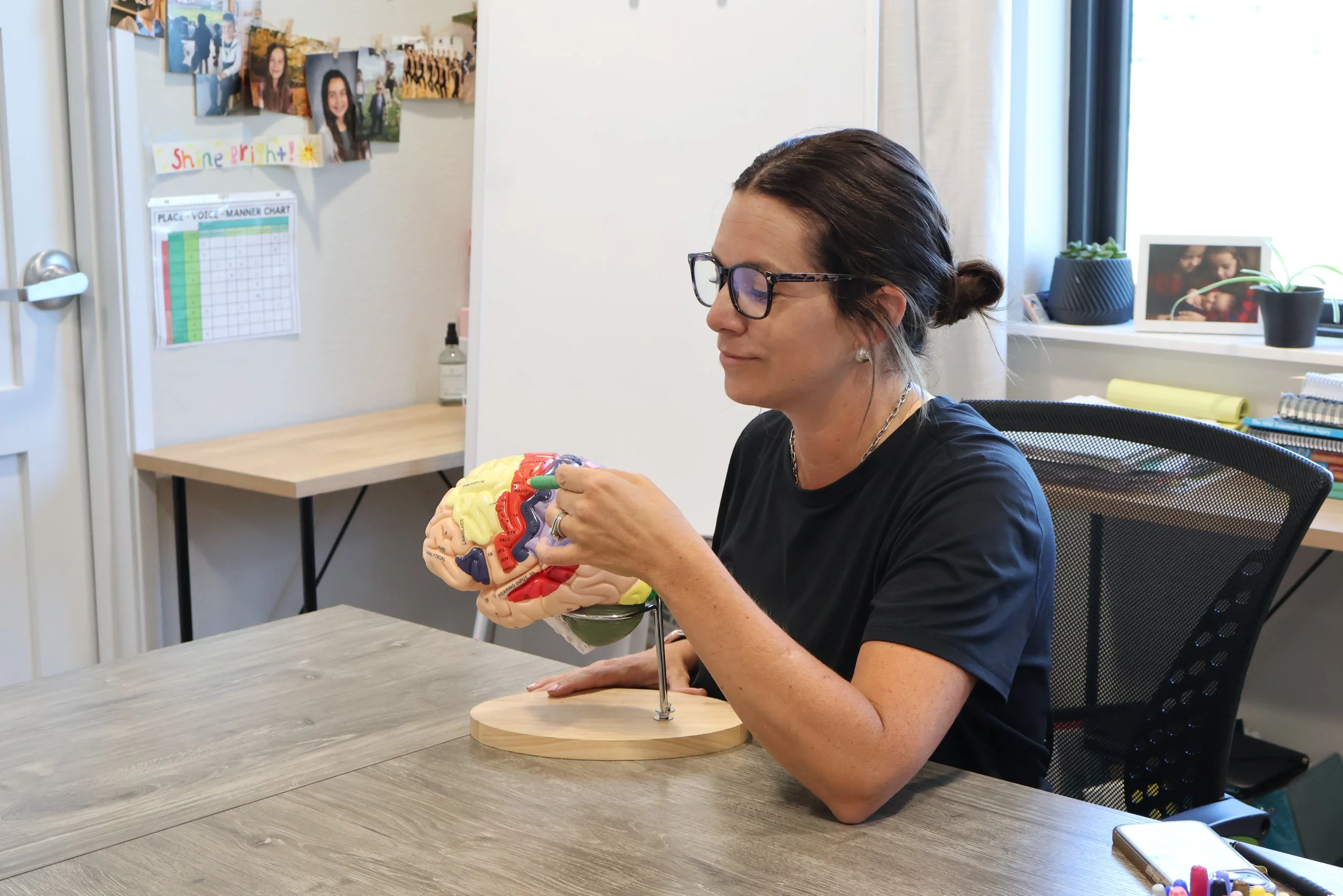 A woman with glasses and dark hair in a bun is sitting at a table, holding a model of a human brain on a stand. The room has photos and artwork on the wall, plants, and a window in the background.