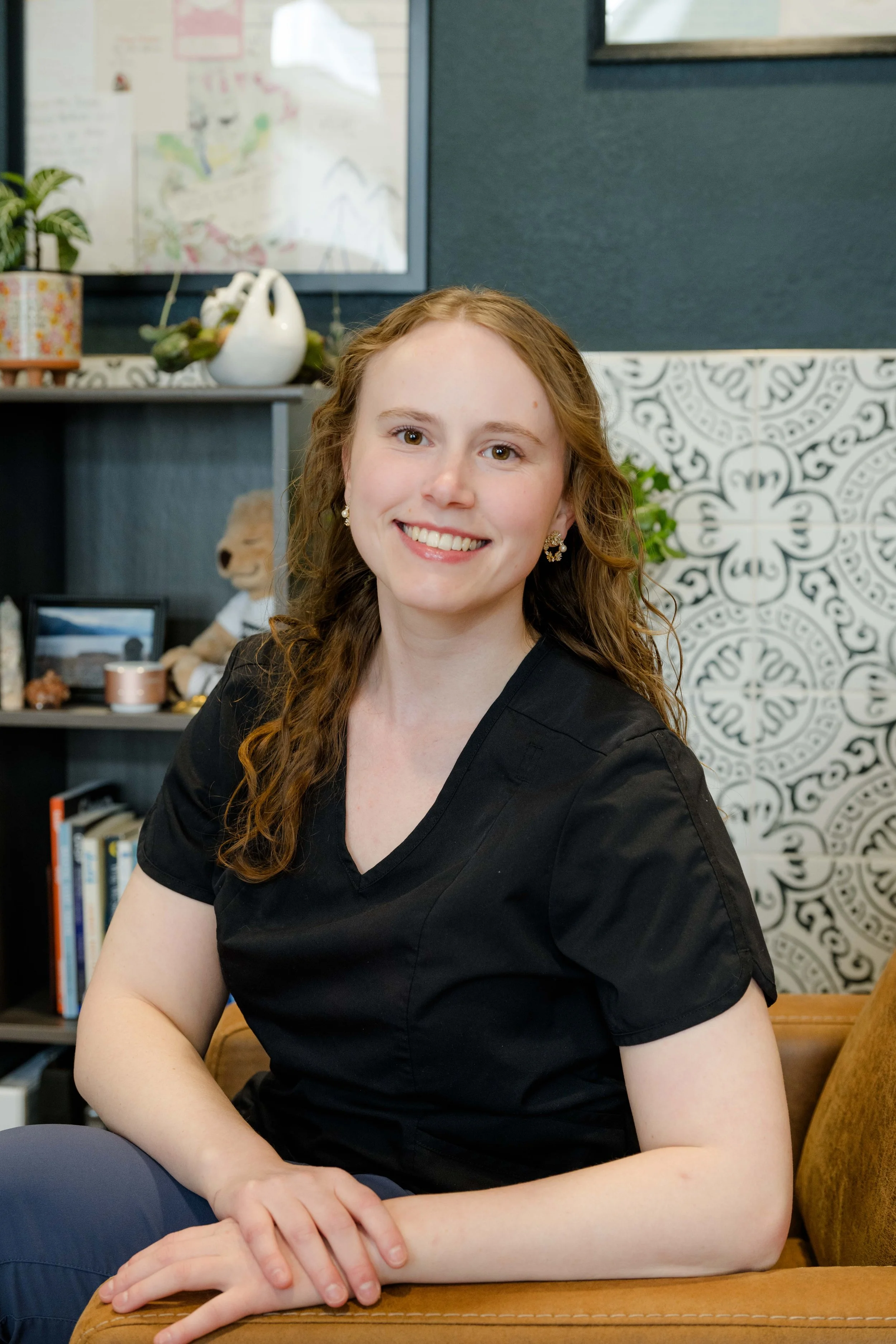 A woman with long, curly reddish hair smiling in an office or living room setting. She is wearing pearl earrings and a black shirt. Behind her, there is a bookshelf with books and decorations, a teddy bear, and a wall with a decorative tile pattern.