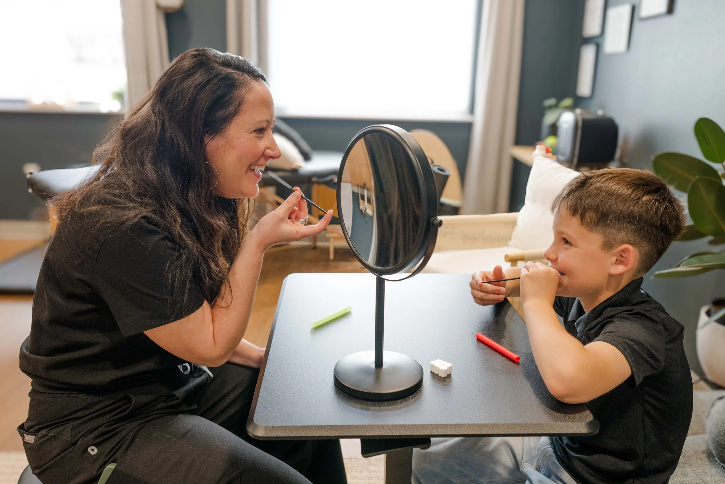 A woman and a young boy sit across from each other at a table with a mirror between them. The woman is holding a makeup brush, smiling, while the boy, also smiling, holds a makeup brush near his chin. There are small objects on the table, and the background shows a room with windows and framed pictures.