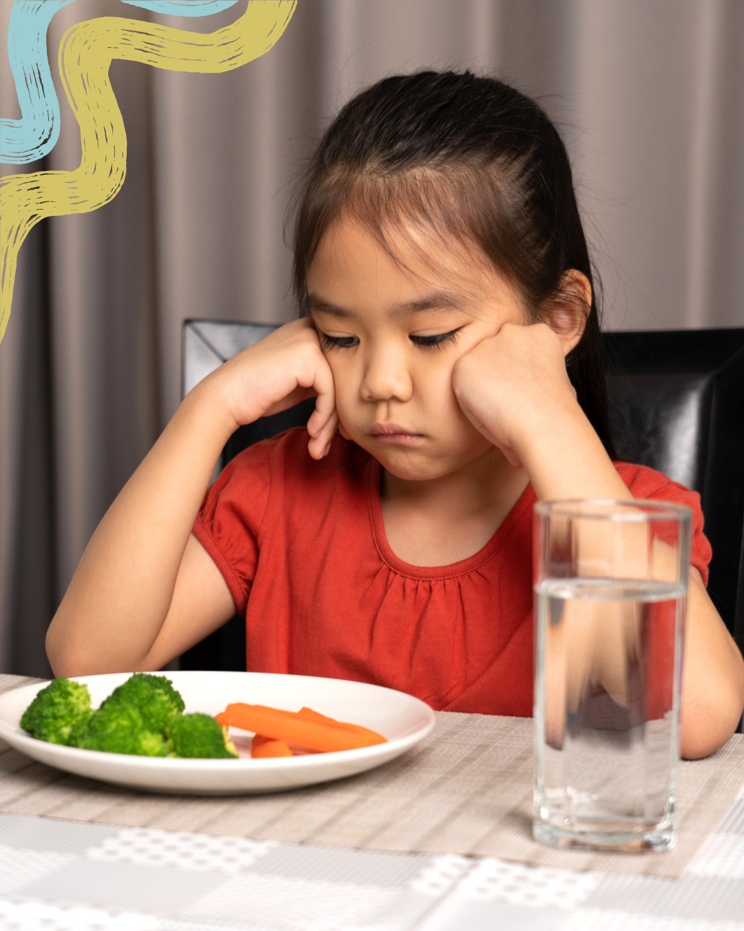 A young girl with a frown, sitting at a dining table with a plate of broccoli and carrots in front of her, and a glass of water nearby.
