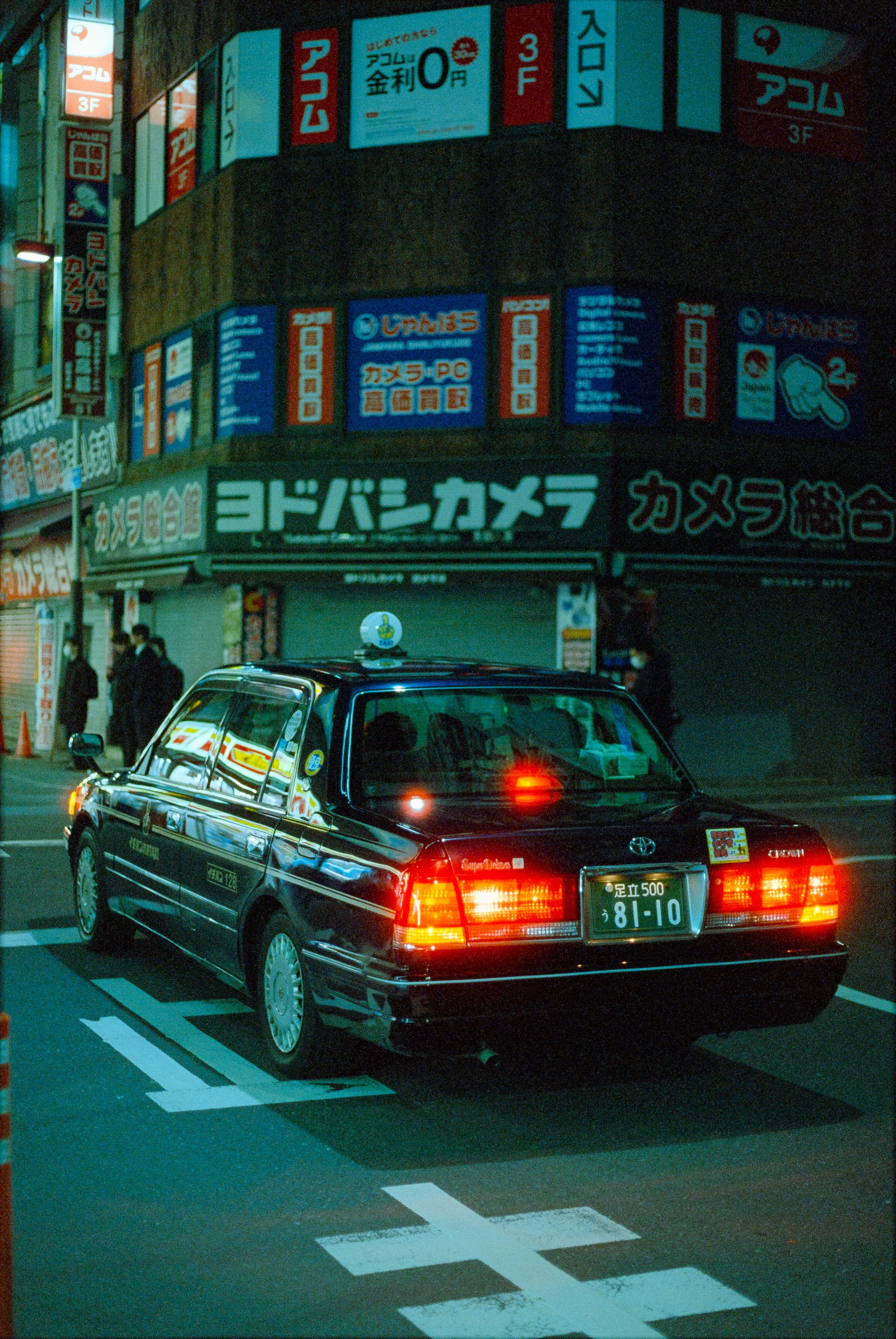 A black taxi cab with Japanese license plates and a taxi sign on the roof, parked on a city street at night with illuminated signs and storefronts in Japanese in the background.