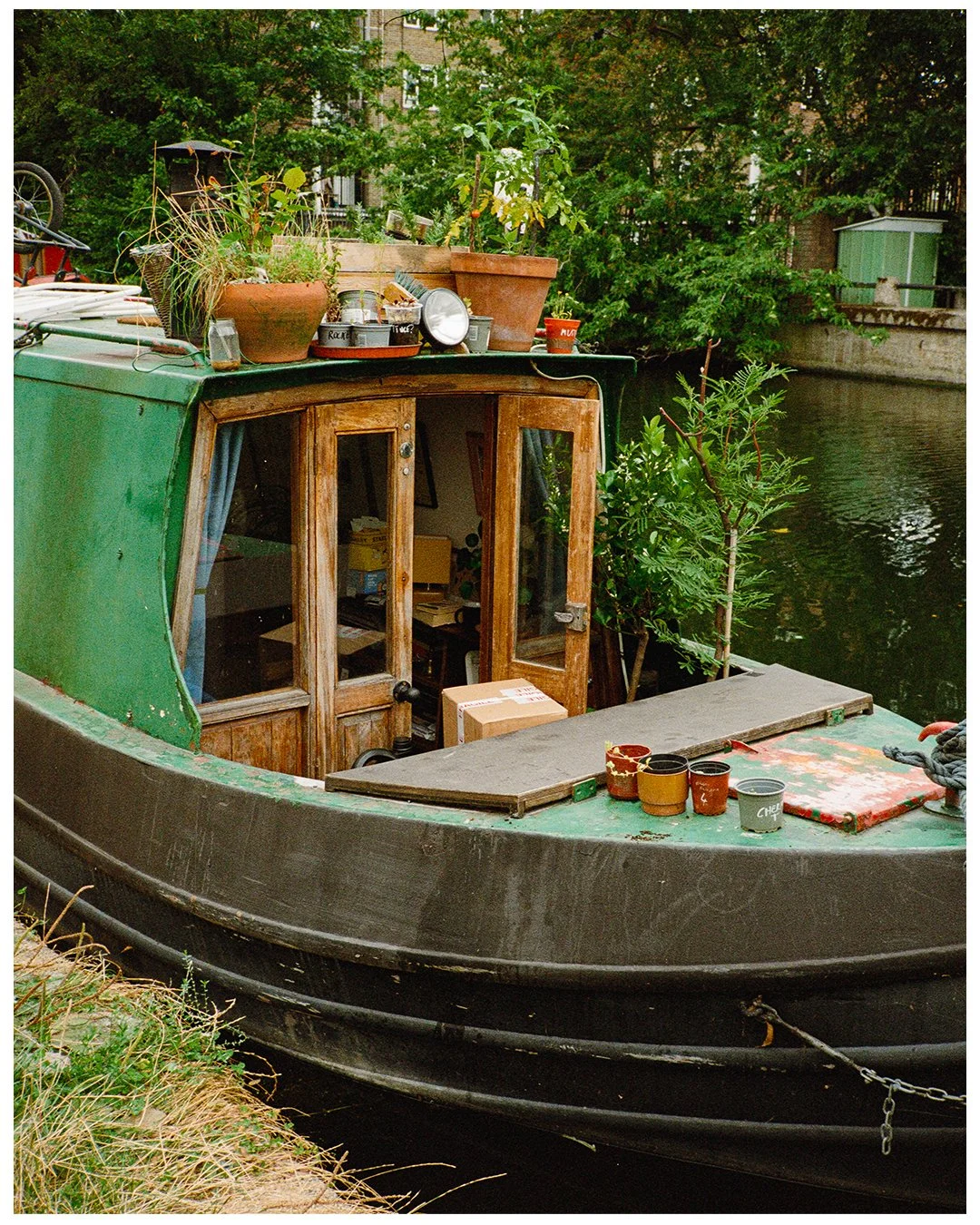 A houseboat with a green exterior and wooden door, docked along a river, with potted plants and gardening supplies on top, surrounded by trees and urban buildings.