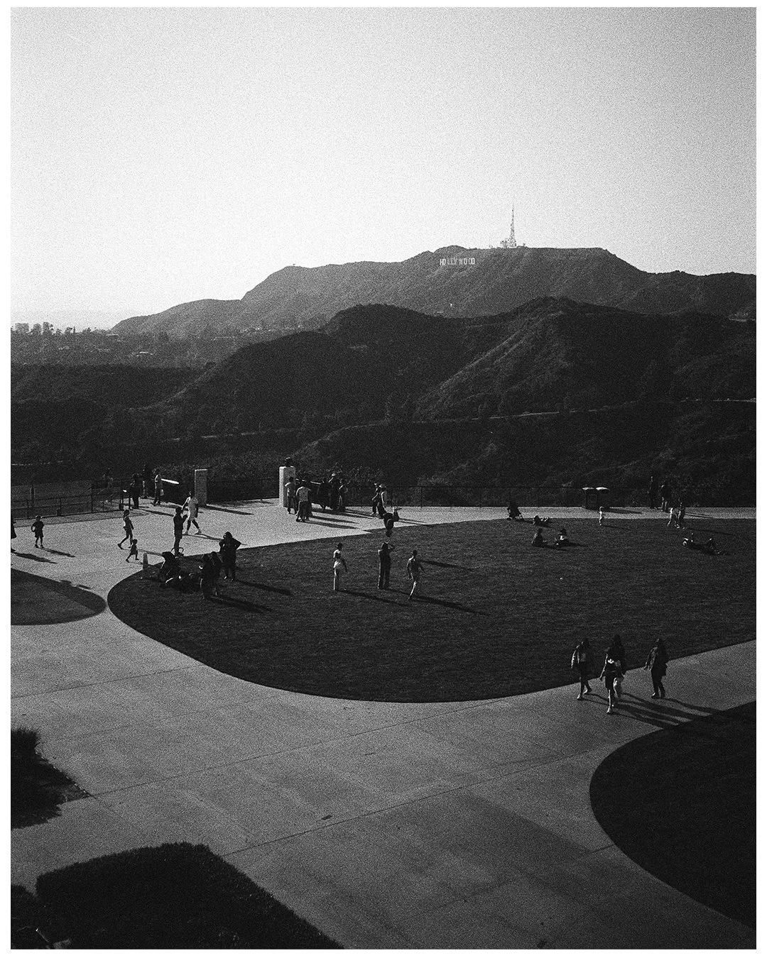 People walking and sitting in a park area with the Hollywood sign visible on the hillside in the background, black and white photo.