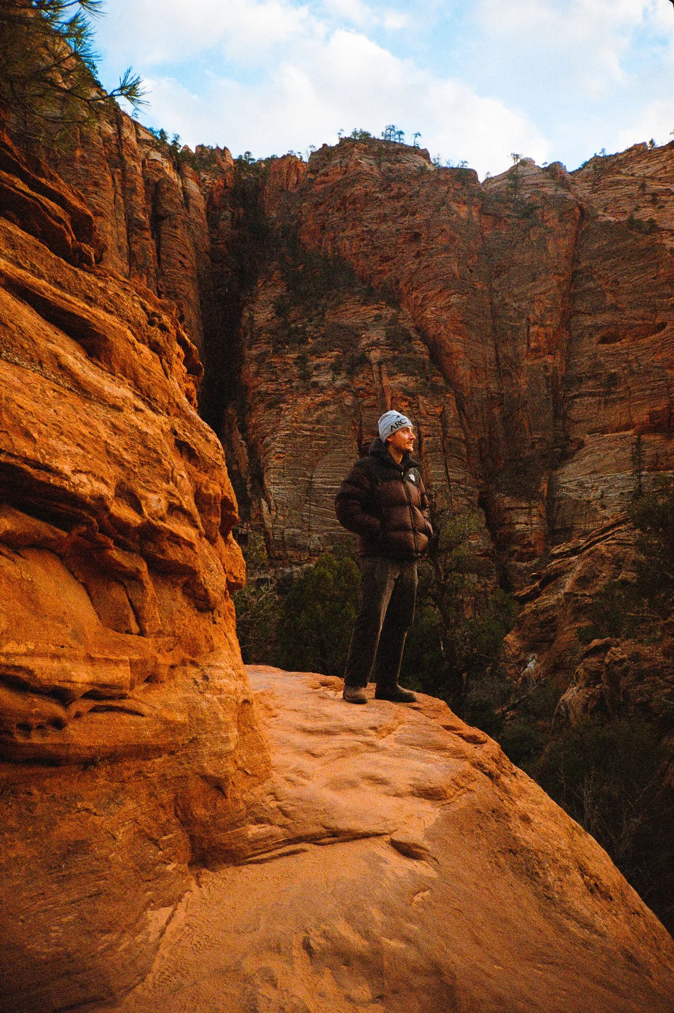 A man standing on a rocky ledge in a canyon, surrounded by towering reddish-orange rock formations, wearing a black puffer jacket and a beanie.