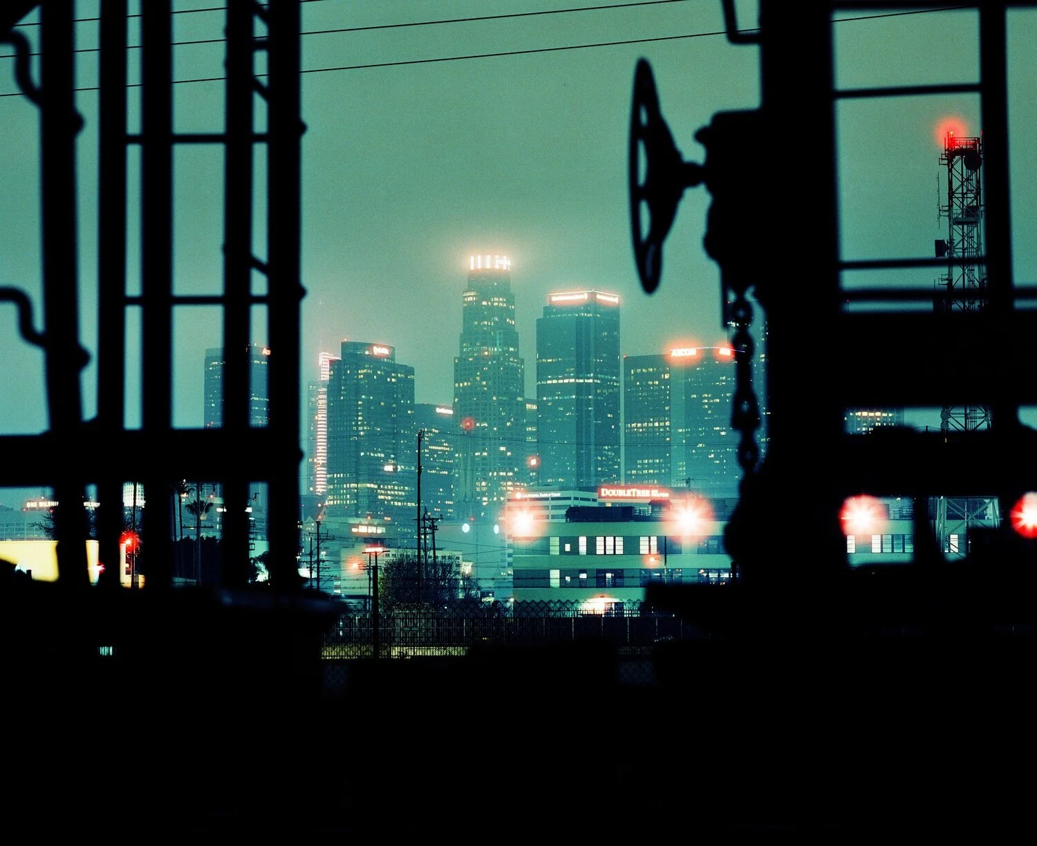 City skyline at night viewed through construction or utility fences with illuminated buildings and overcast sky.